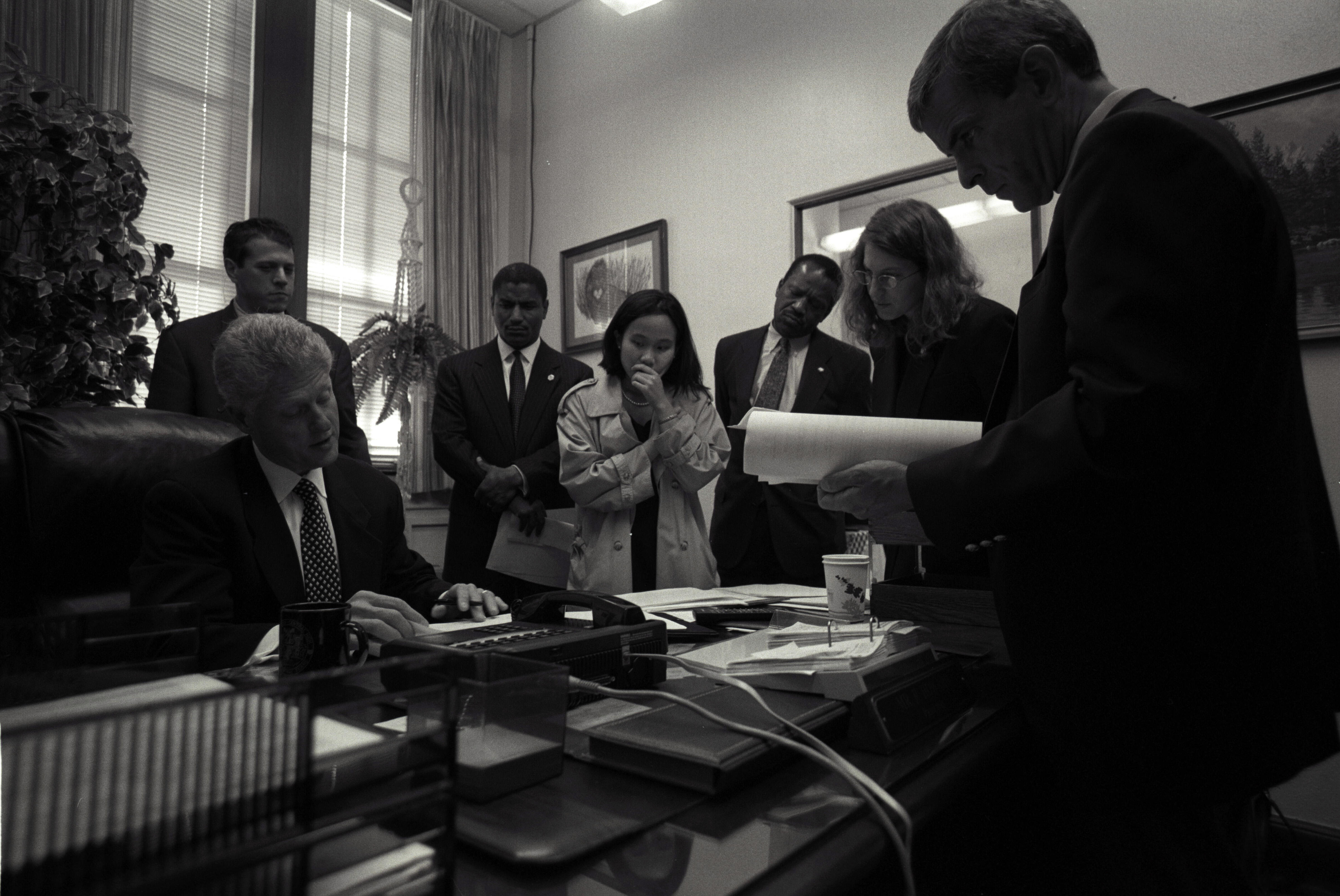 Black and white photograph. President Clinton sits at a desk, others are standing.