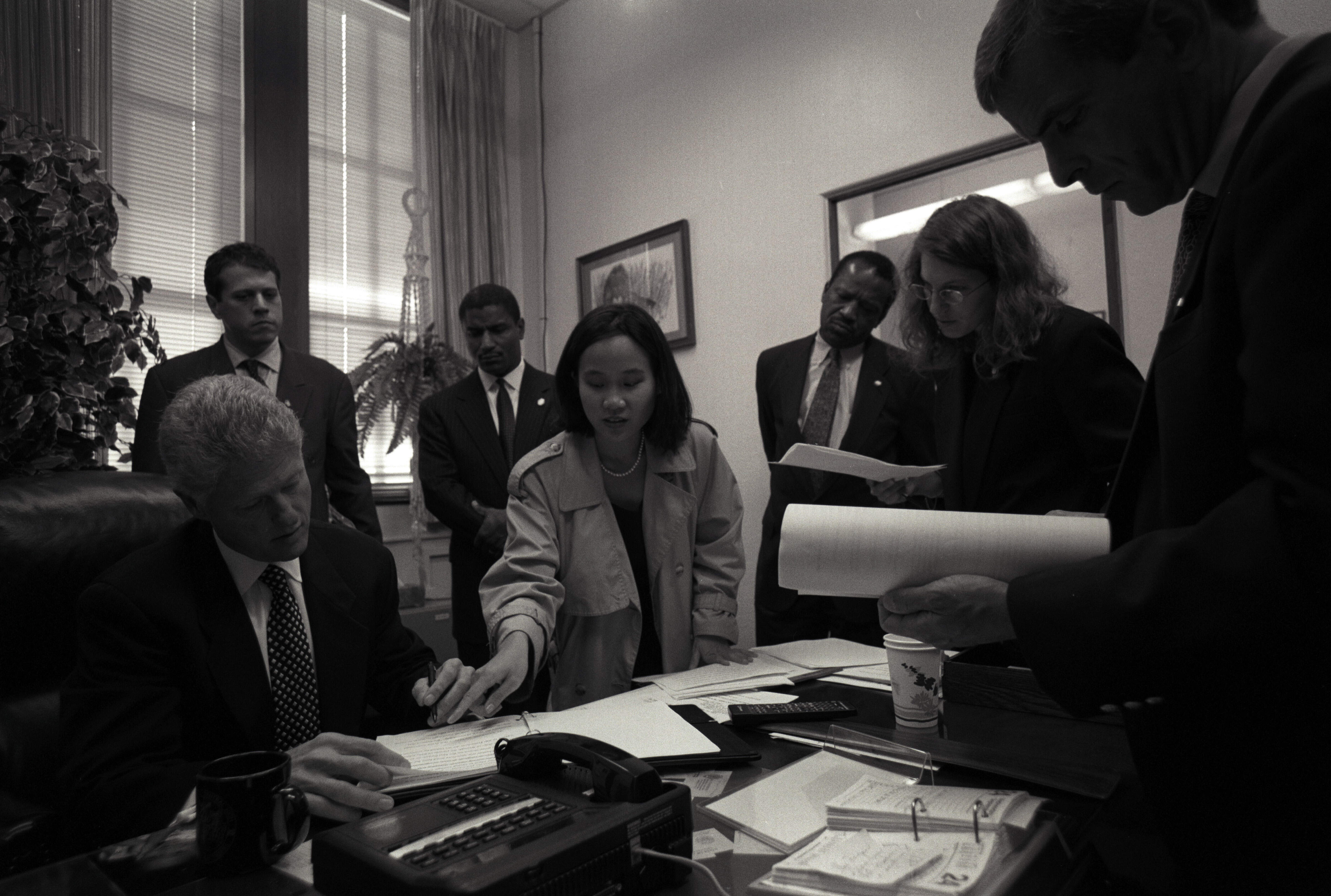 Black and white photograph. President Clinton sits at a desk. Speechwriter, June Shih leans over the desk to point at speech draft, President Clinton is looking down at the paper.