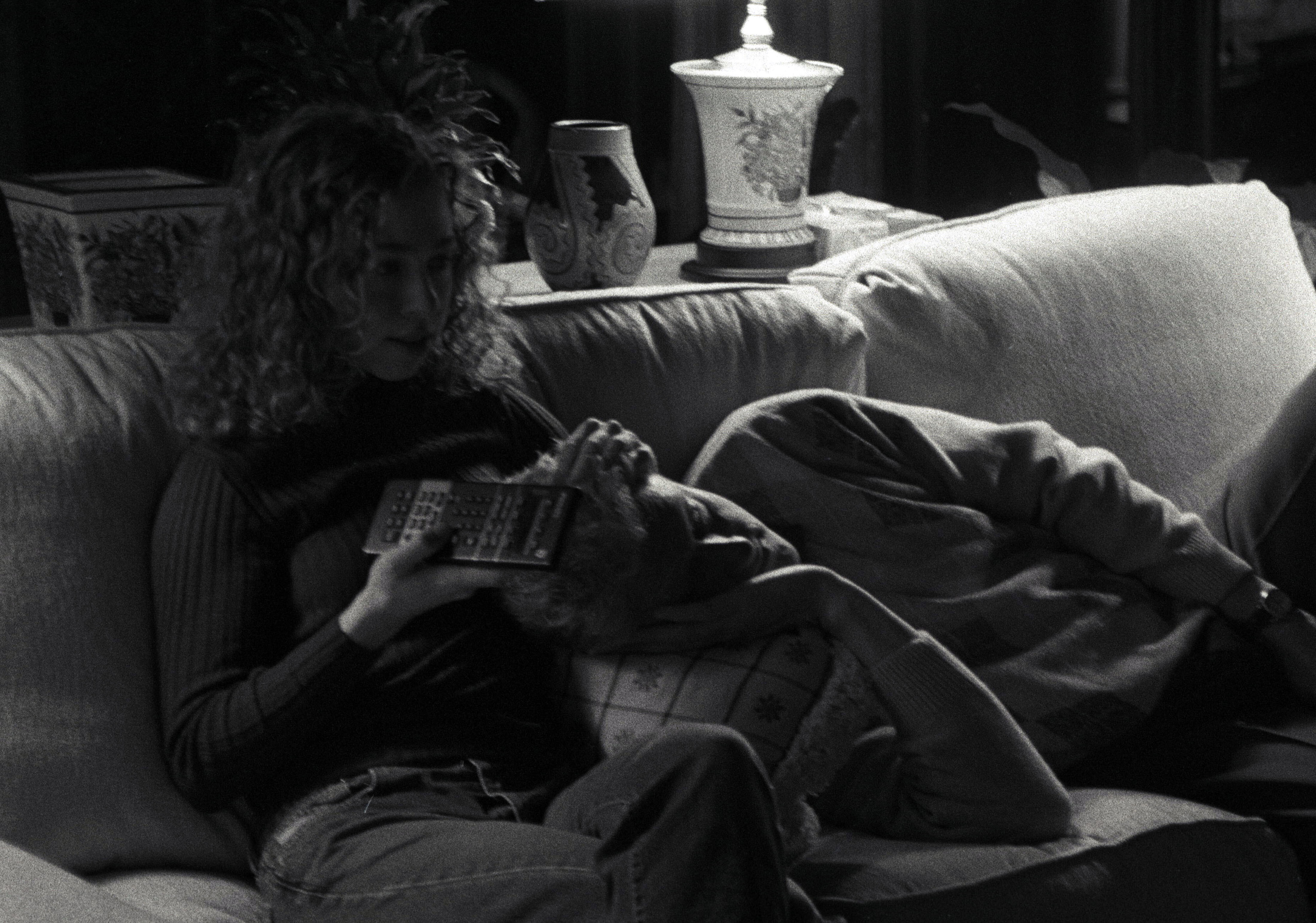 Black and white photograph. President Clinton reclines on a couch, with his head on a pillow, next to his daughter, Chelsea Clinton as she holds a television remote control.