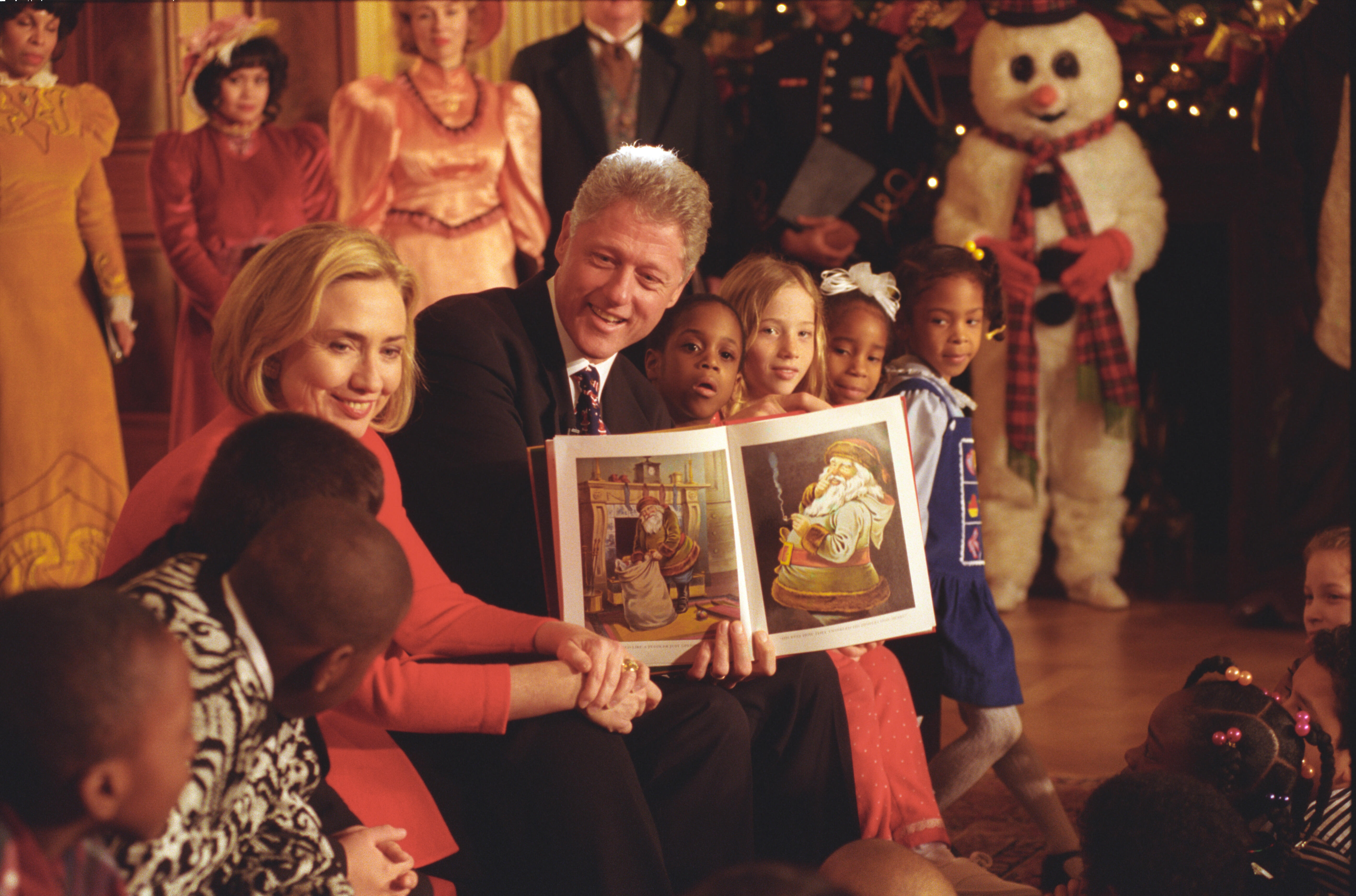 President Clinton and First Lady Hillary Rodham Clinton sit and hold up the book to show the book's illustrations to children sitting around them. People in period costumes can be seen standing in the back ground. a person dressed as a snow man can also be seen in the back ground.