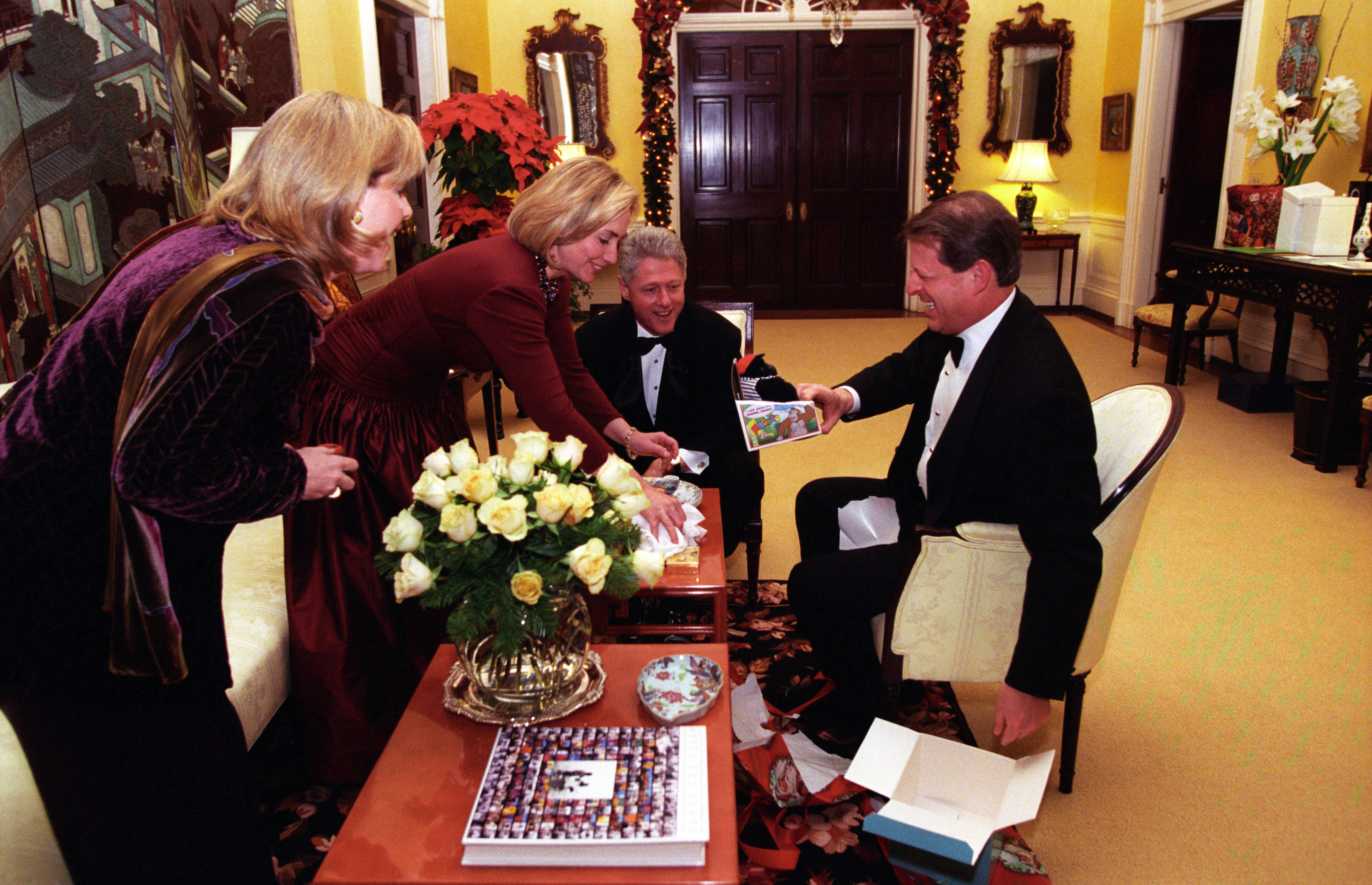 President Clinton and Vice President Al Gore sit in white chairs. First Lady Hillary Rodham Clinton and Tipper Gore stand leaning over a table, all look at a Christmas gift in Vice President Gore's hand.