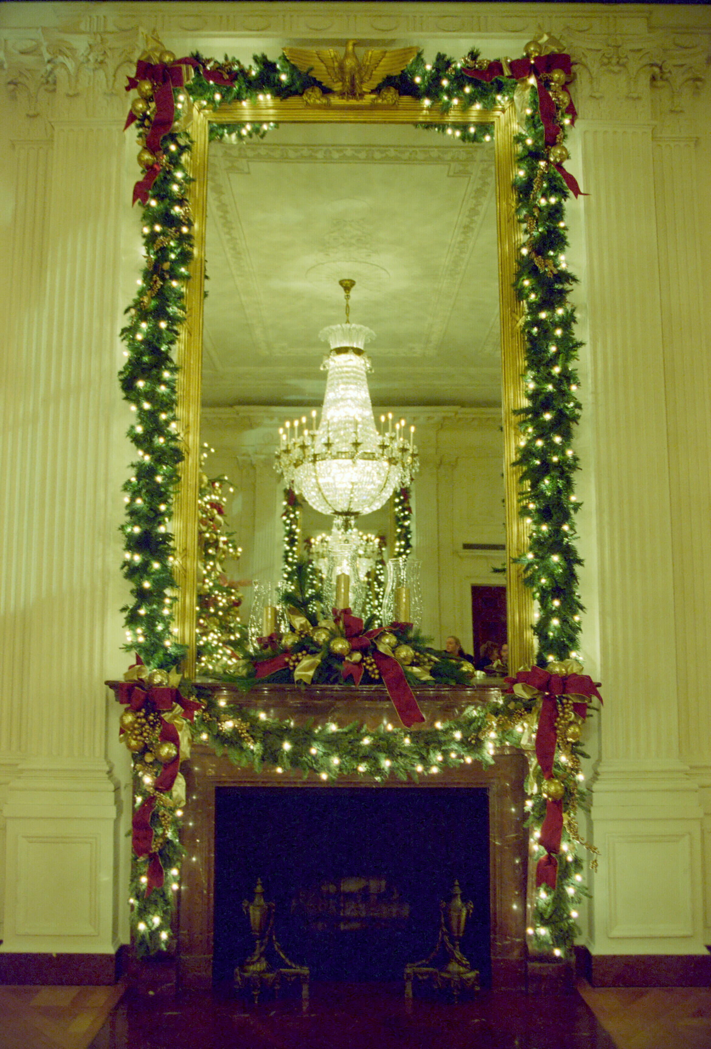 Christmas decorations on the fireplace mantel. Lit garland surrounds the mantle from the floor to ceiling. A reflection of a chandelier can be seen in a mirror above the mantel.