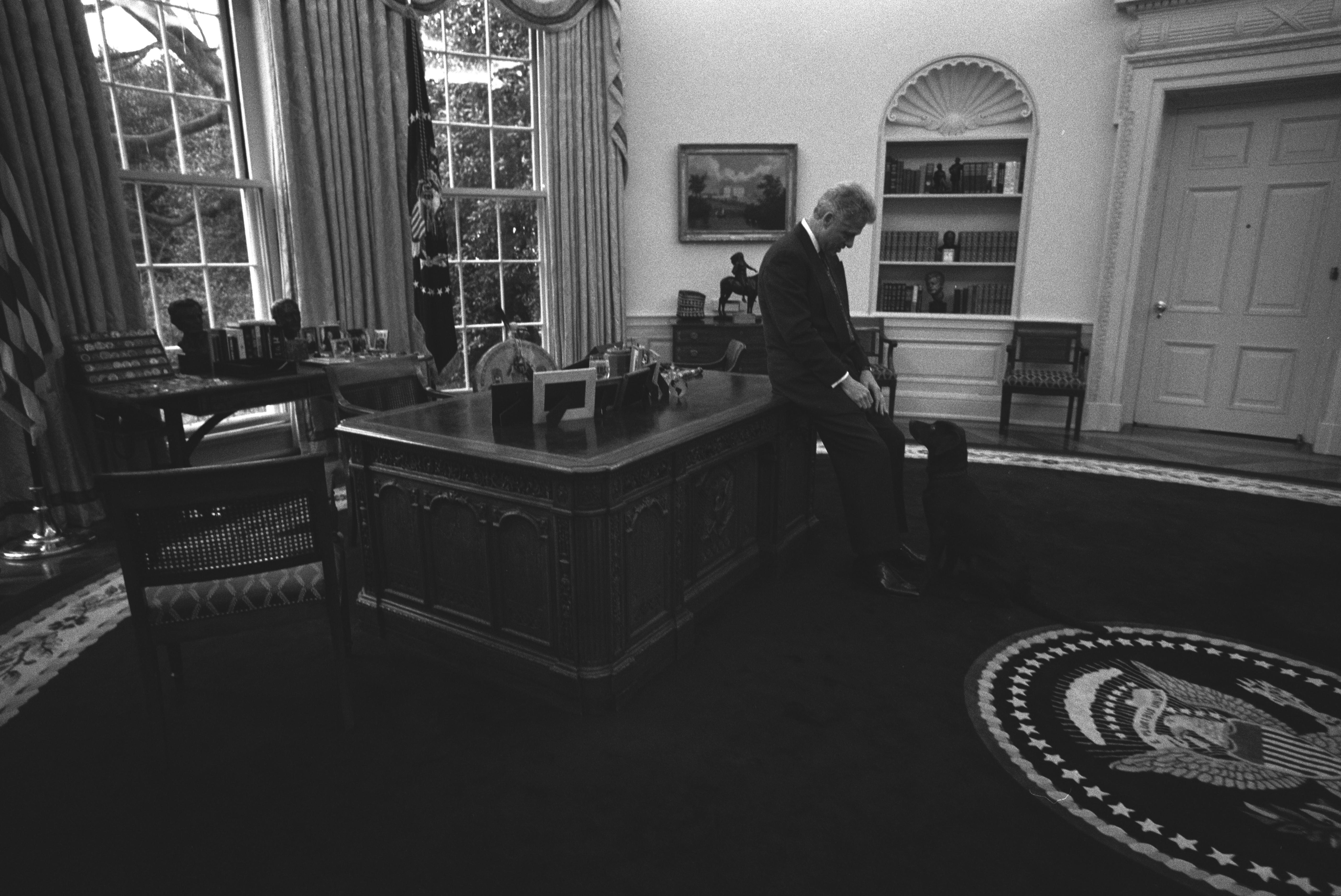 Black and white photograph. President Clinton sits on the edge of the Resolute Desk and plays with Buddy the Dog in the Oval Office following a briefing prior to a Radio Address