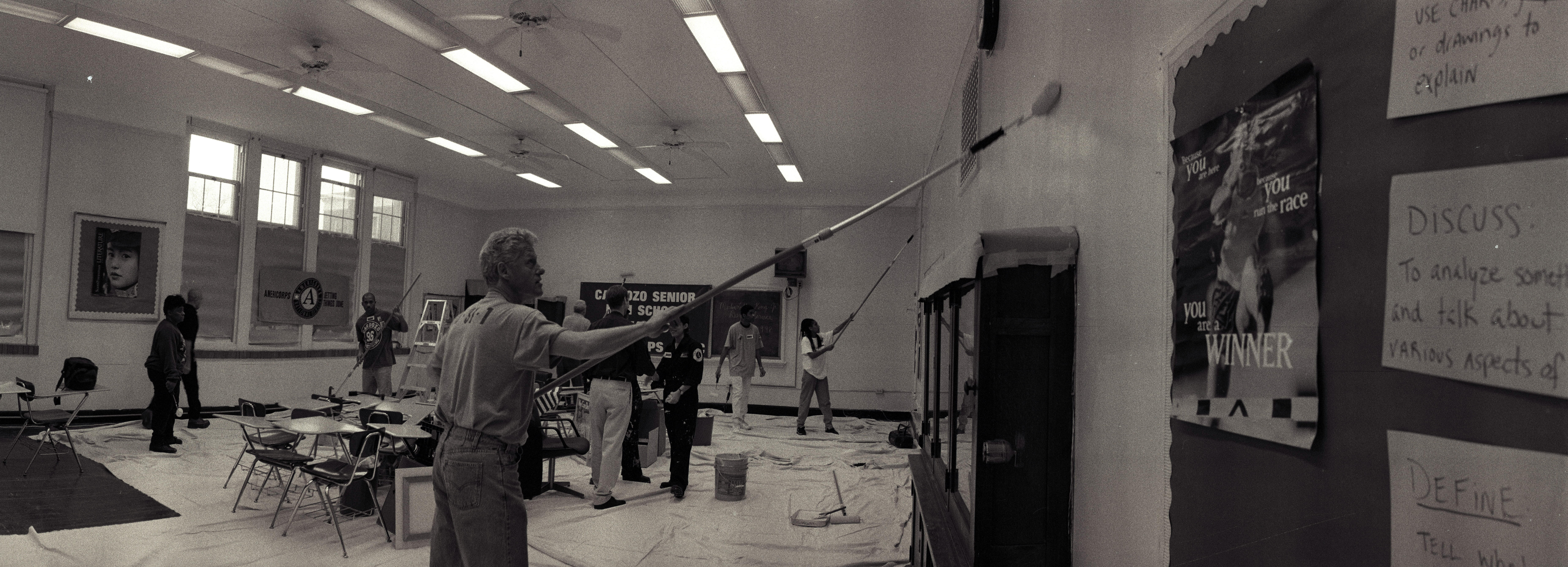 Black and white photograph. President Clinton is casually dressed in a t shirt and jeans. President Clinton uses a long handed paint roller to pain the walls of a classroom. others are also painting and working in the background