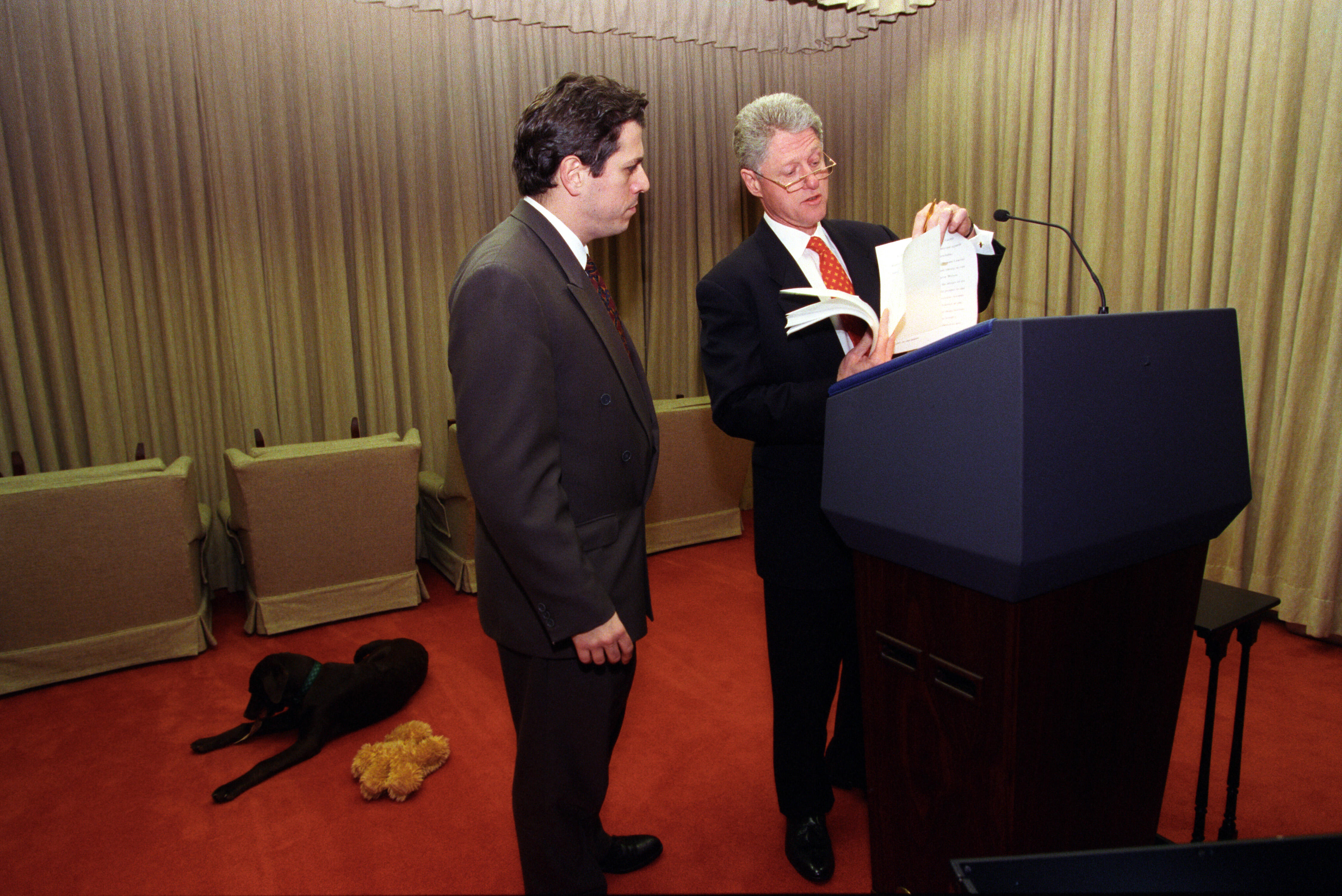 President Clinton works on his speech in the Family Theatre. Buddy the dog lays on the floor with a soft toy.The President stands with a speechwriter behind a podium, wearing glasses, holding a speech draft.
