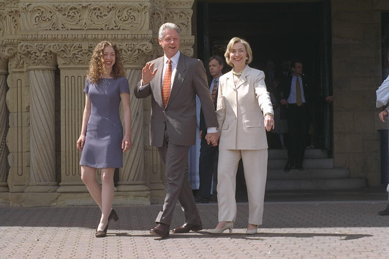 President Clinton, Hillary Rodham Clinton, and Chelsea Clinton depart church services at Stanford University Memorial Church. President Clinton waves at the press with his right hand and holds Hillary Rodham Clinton's hand with his left hand.