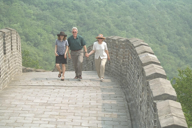 President Clinton, Hillary Rodham Clinton, and Chelsea Clinton hold hands as they walk along the Great Wall. The First Lady and Chelsea wear hats and sunglasses.