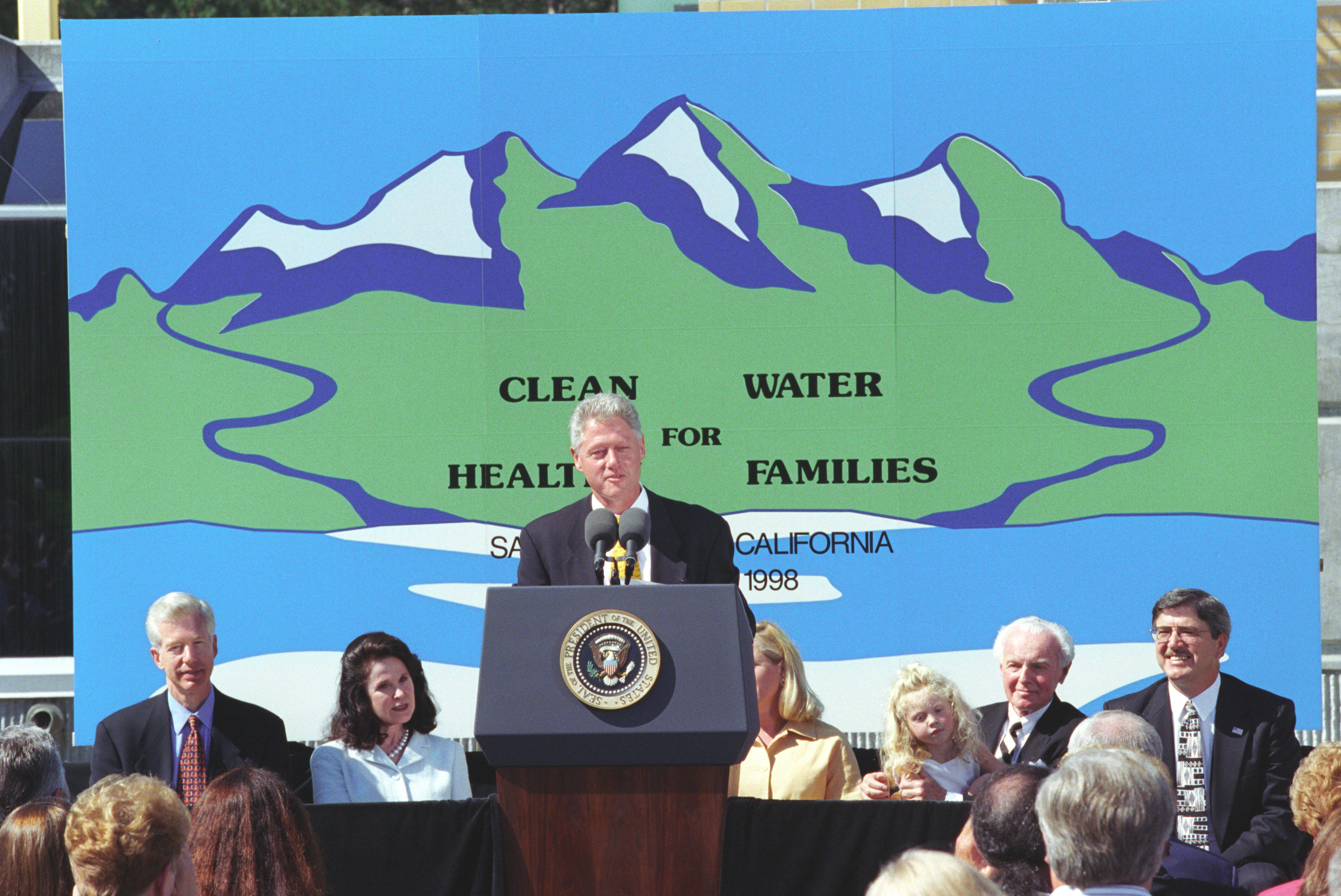 President Clinton delivers remarks behind a podium. Behind President Clinton is a large hand painted sign that reads " Clean Water for Healthy Families." The hand painted sign also has an image of snow covered mountains and water. 