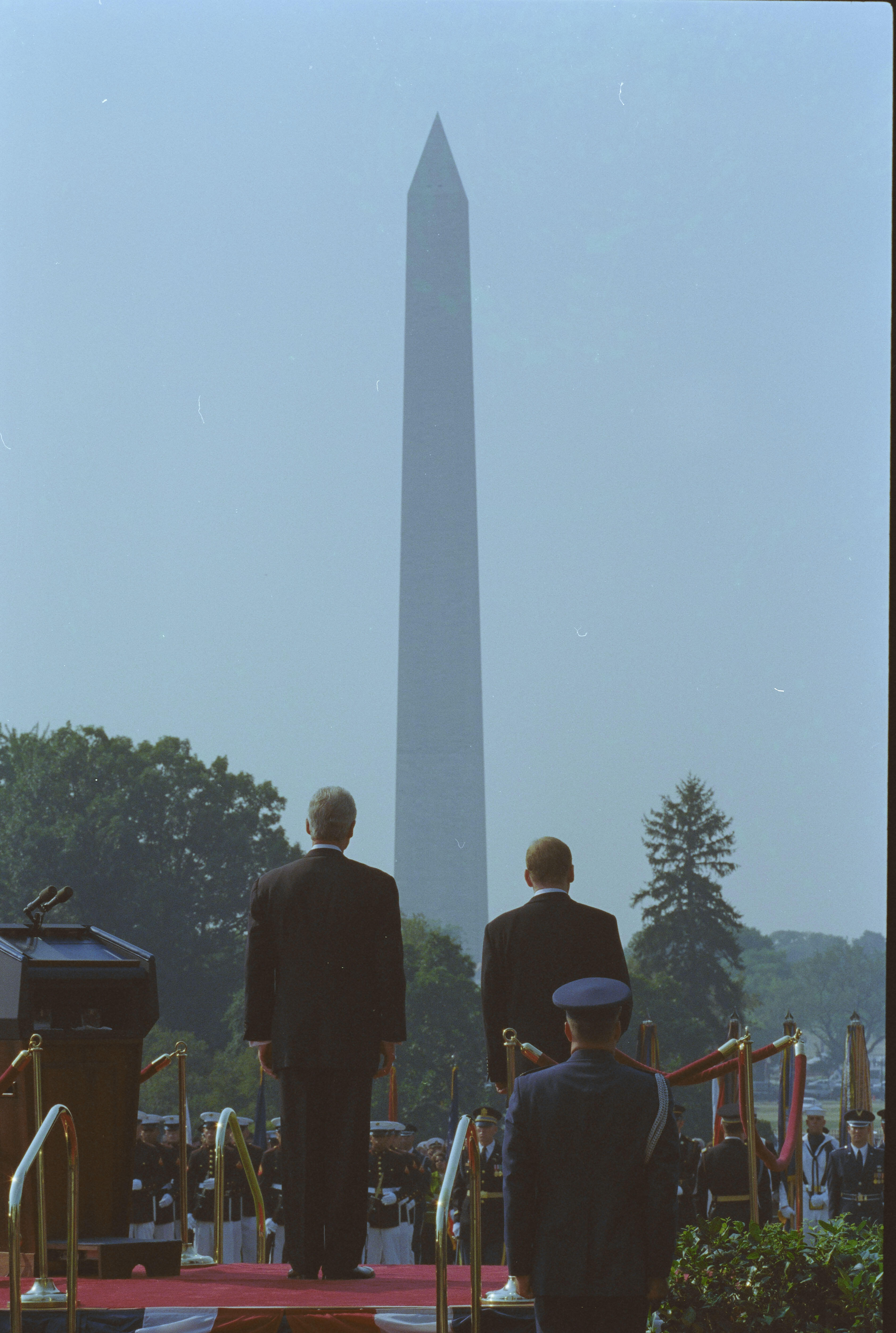 President Clinton and President Václav Havel of the Czech Republic stand on the dais on the South Lawn of the White House looking toward the Washington Monument. Trees can also be see in the background. A man in a uniform is below the dias, also looking toward the Washington Monument (an obelisk)