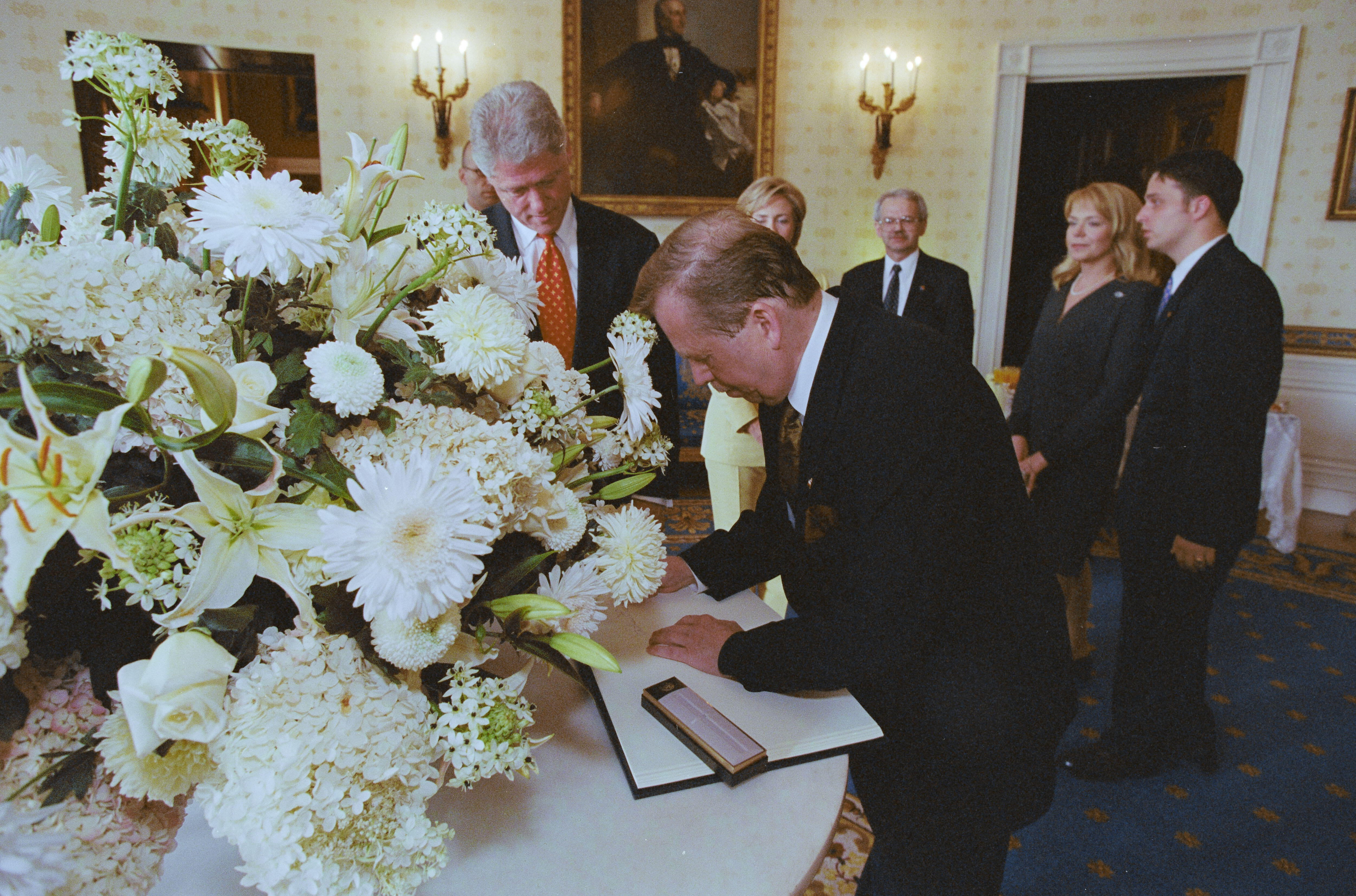 President Václav Havel signs a book with white pages. A pen box sits on the open guest book. The book sits on a round table with a white table cloth. A large flower arrangement of white flowers sits on the table. President Clinton stands looking down at President Havel as he signs the guest book. First Lady Hillary Rodham Clinton and President Havel's wife Dagmar Havlova can be seen standing in the room. Two unidentified men stand in the background.