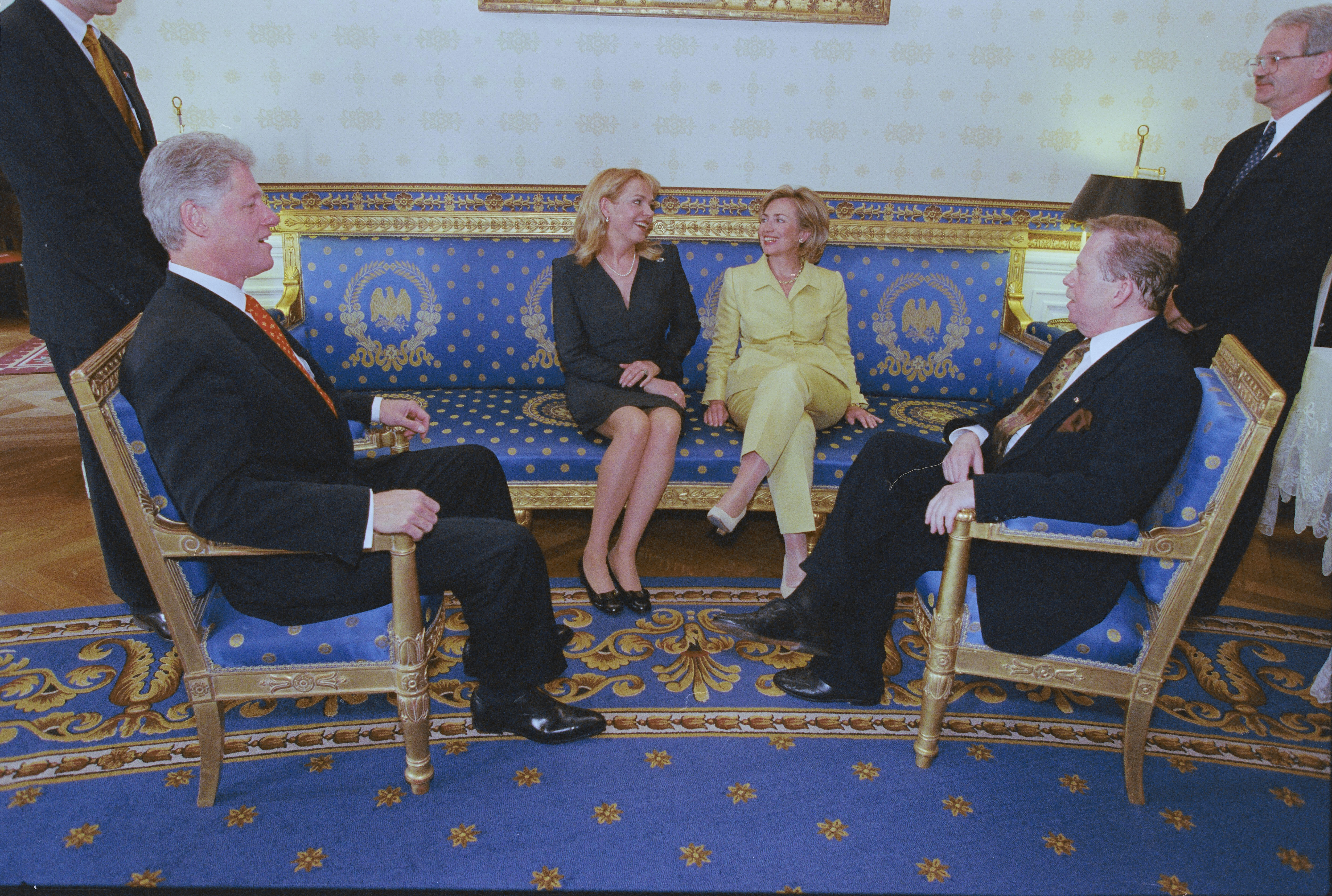 President Clinton, President Václav Havel of the Czech Republic sit across from each other in gold and blue chairs. First Lady Hillary Rodham Clinton, and Dagmar Havlova sit next to each other on a gold and blue bench. They are flanked to the left and right by two unknown men. The room has a large blue capet on light brown wood floors.