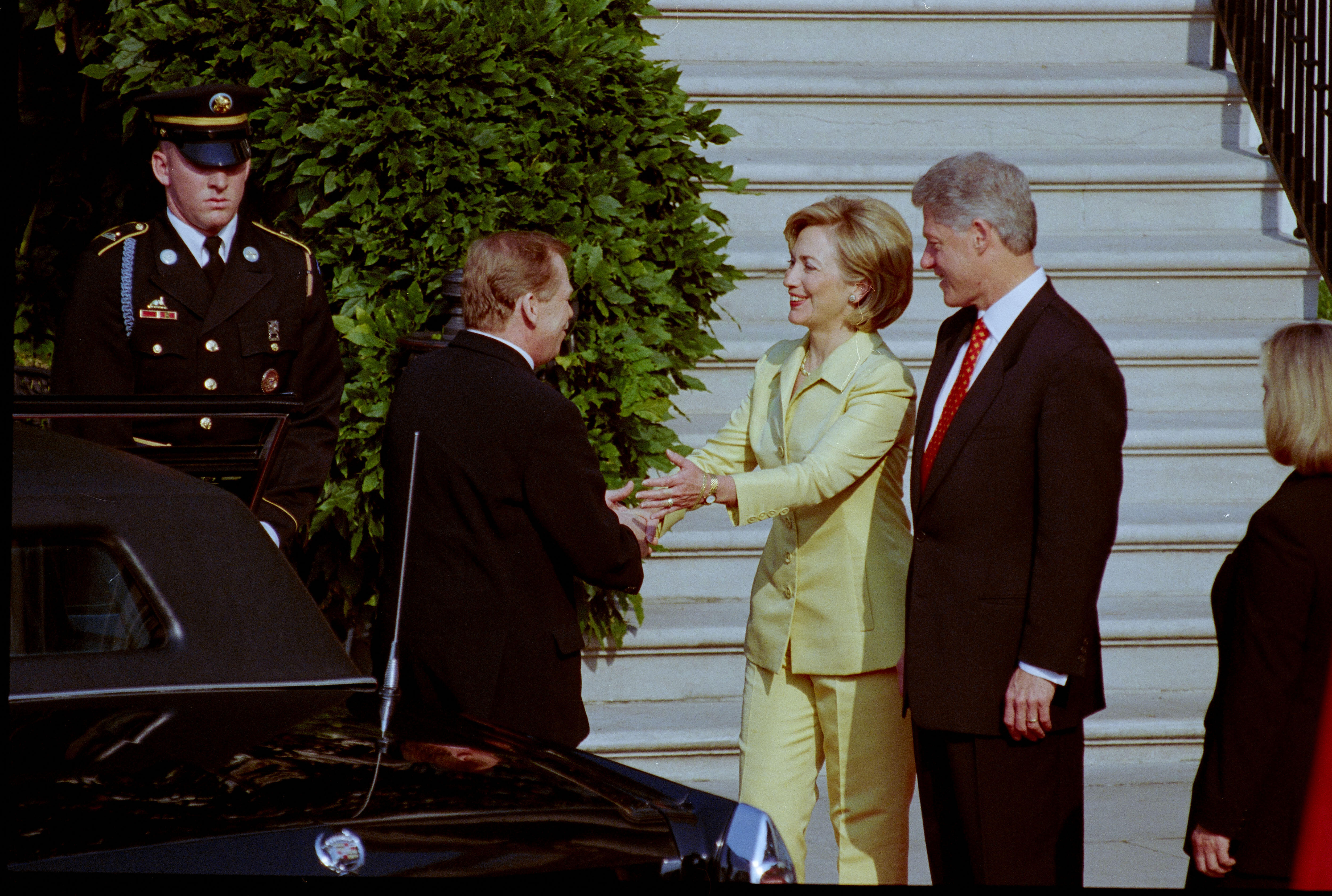 First Lady Hillary Rodham Clinton stands greeting President Václav Havel. First Lady Hillary Rodham Clinton extends both hands towards Havel. She wears a yellow pantsuit. President Clinton stands looking on. President Havel stands beside a black car. A uinformed soldire stands in the background. A green bush and stairs are also in the background. An unknow woman stands lookiing toward the the Clintons and Havel.