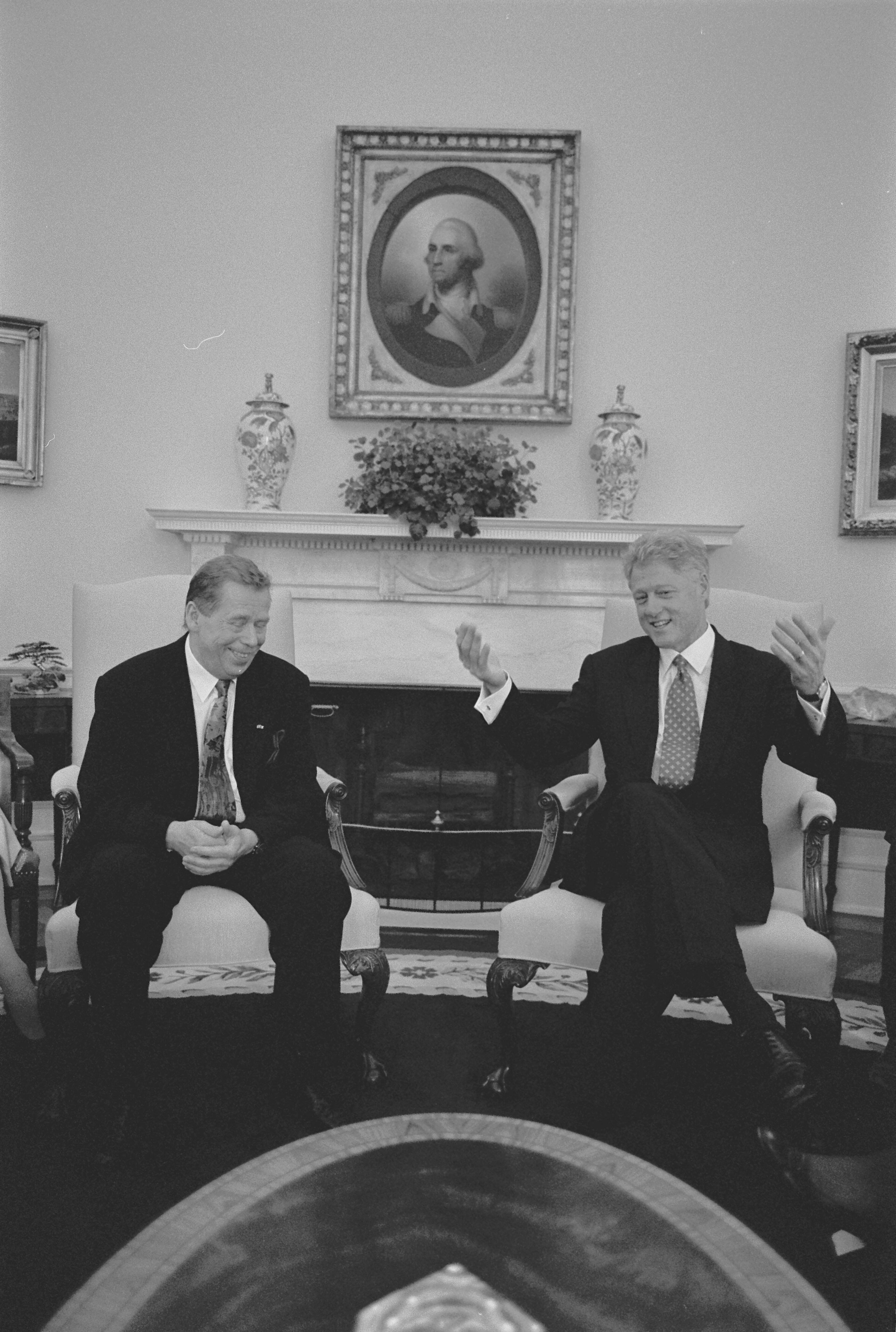 Black and White photograph. President Václav Havel of the Czech Republic sit next to each other in arm chairs in the Oval Office. President Clinton Holds his arms and hands out in a gesture. President Havel looks down, smiling with his hands folded in front of him. Hanging above the mantel is a famous "porthole" portrait of George Washington by artist Rembrandt Peale.