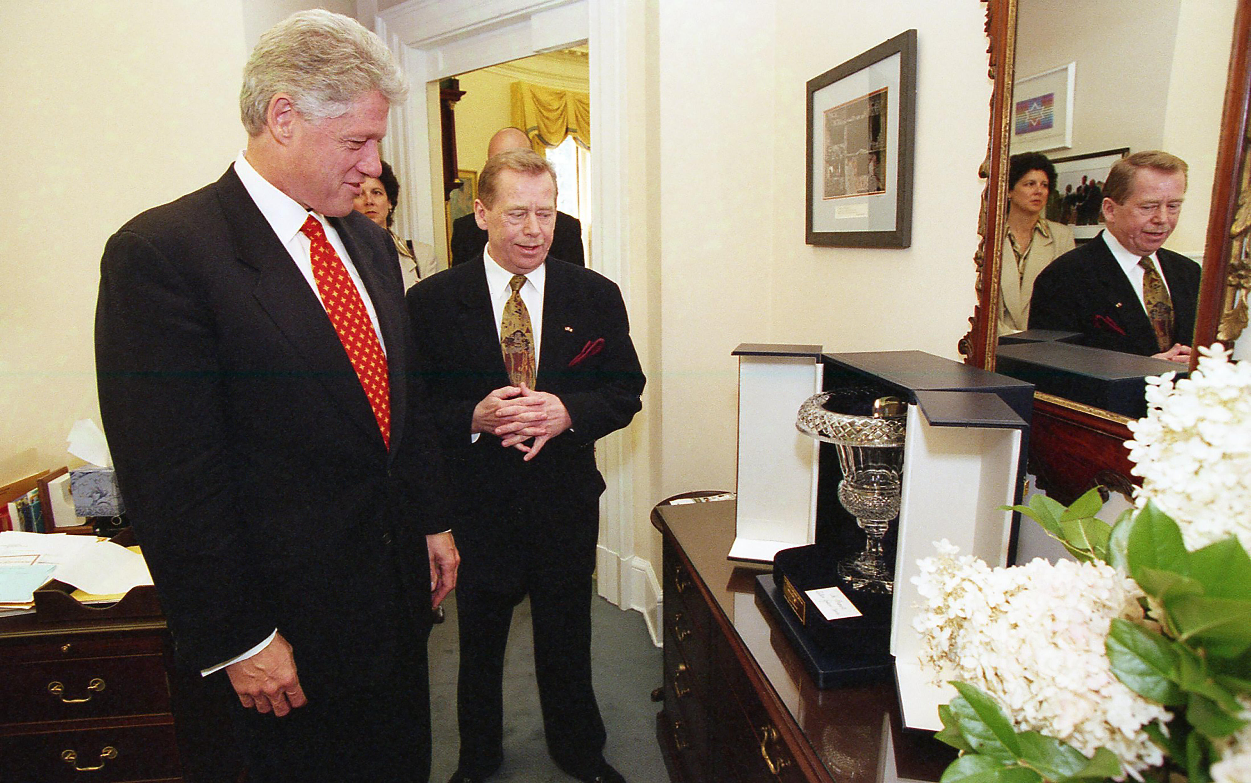 Presdint Clinton stands looking down at aa large crystal vase in a blue leather case sitting on a table. President Václav Havel also looks down at the git on the table. Havel holds his hands in front of him, fingers folded.  White hydrangea flowers sit next to the vase. A mirror on the wall feflects the image of Havel and an unidentified woman.