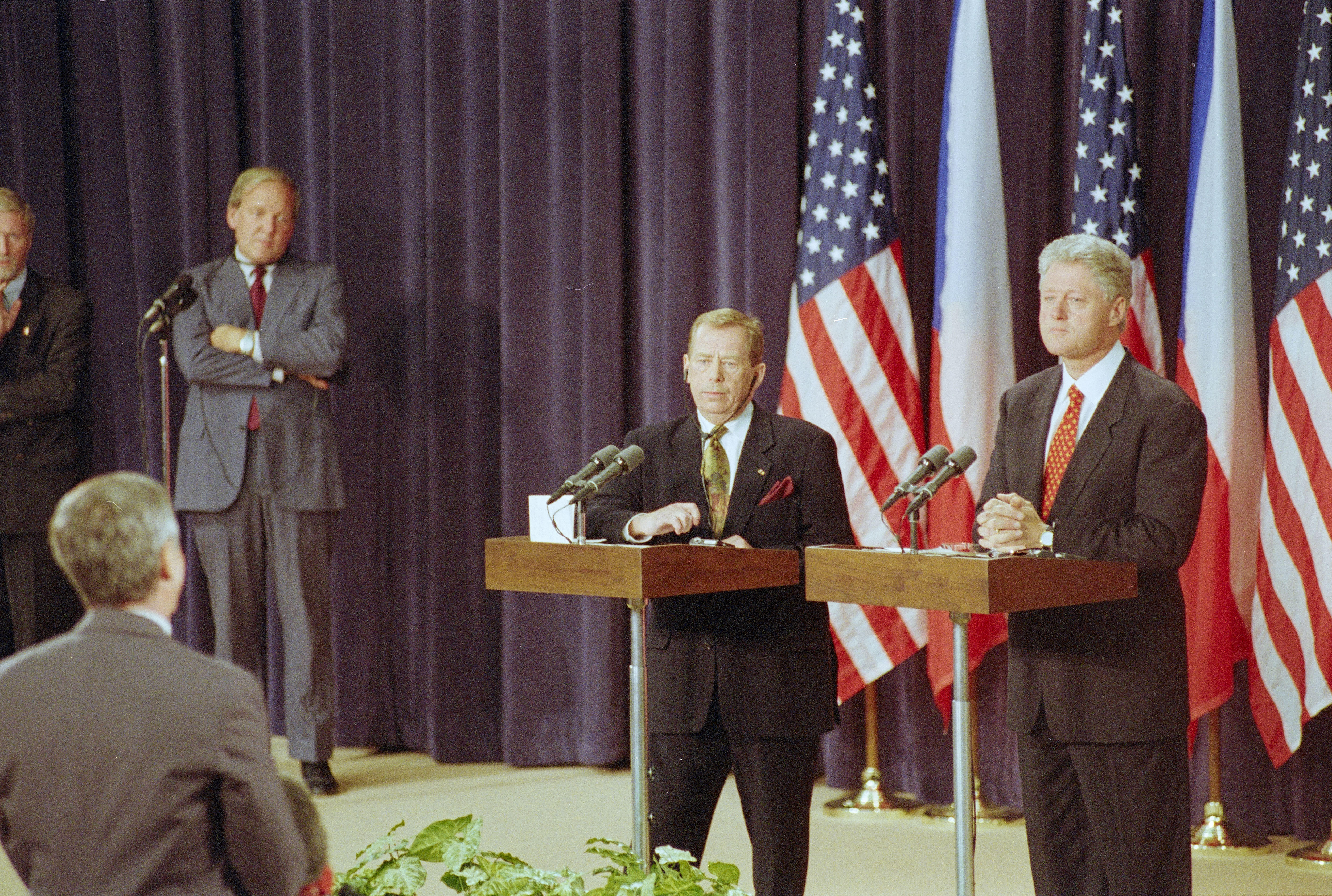 President Clinton and President Václav Havel stand a podiums facing journalists.  Three United States flags and two Czech republic flags are in the background. Two unidentified men can be seen standing behind the presidents (left). A man is standing in front of the Presidents looking toward the Presidents.