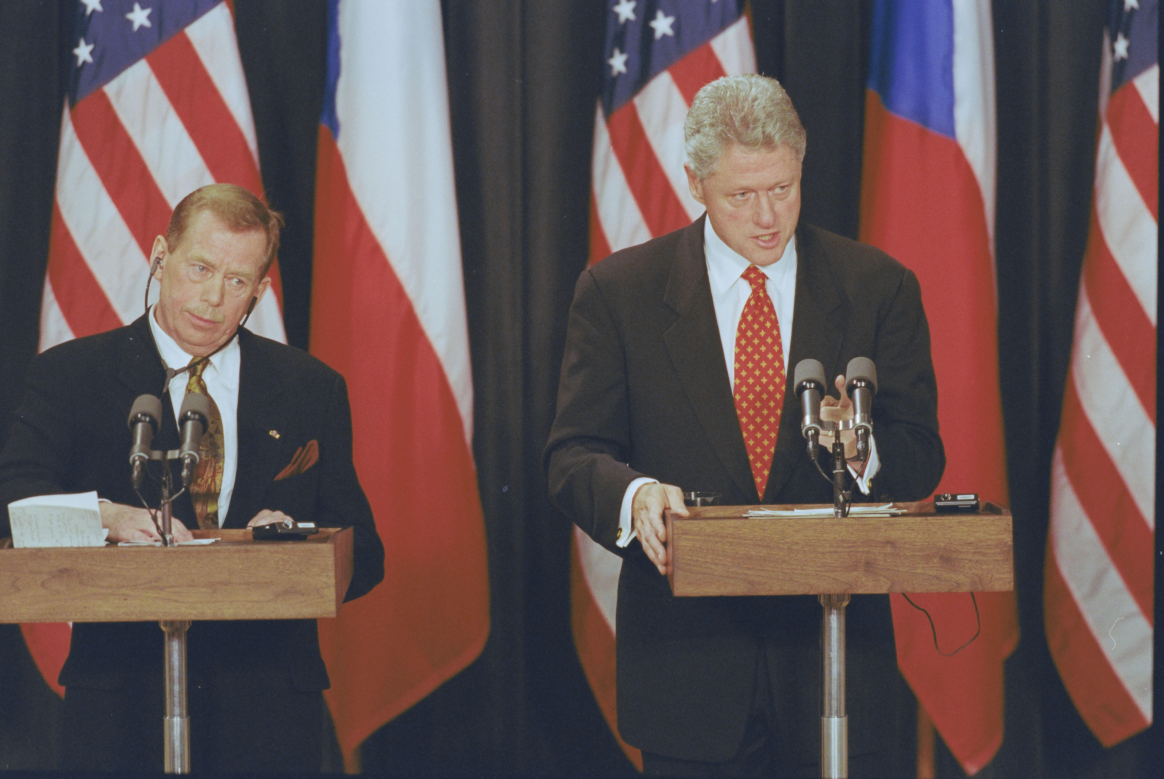President Clinton and President Václav Havel stand behind podiums. President Havel wears erphone headset for translation. Bhind them are three United States flags and two Czech Republic flags.  
