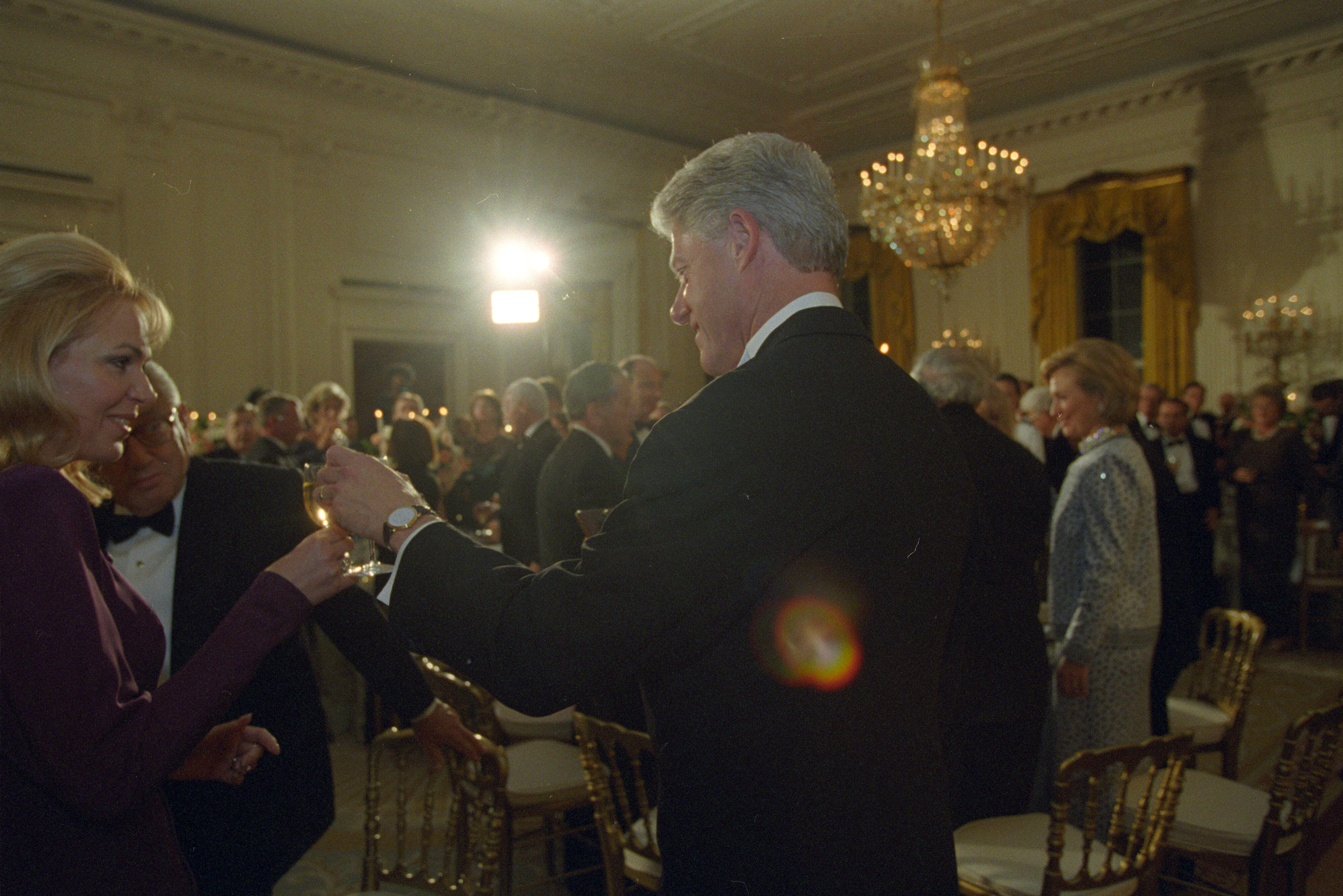 President Clinton stands and touches glasses with Dagmar Havlová. Other people can be seen standing around tables toasting glasses in the State Dining room of the White House. 