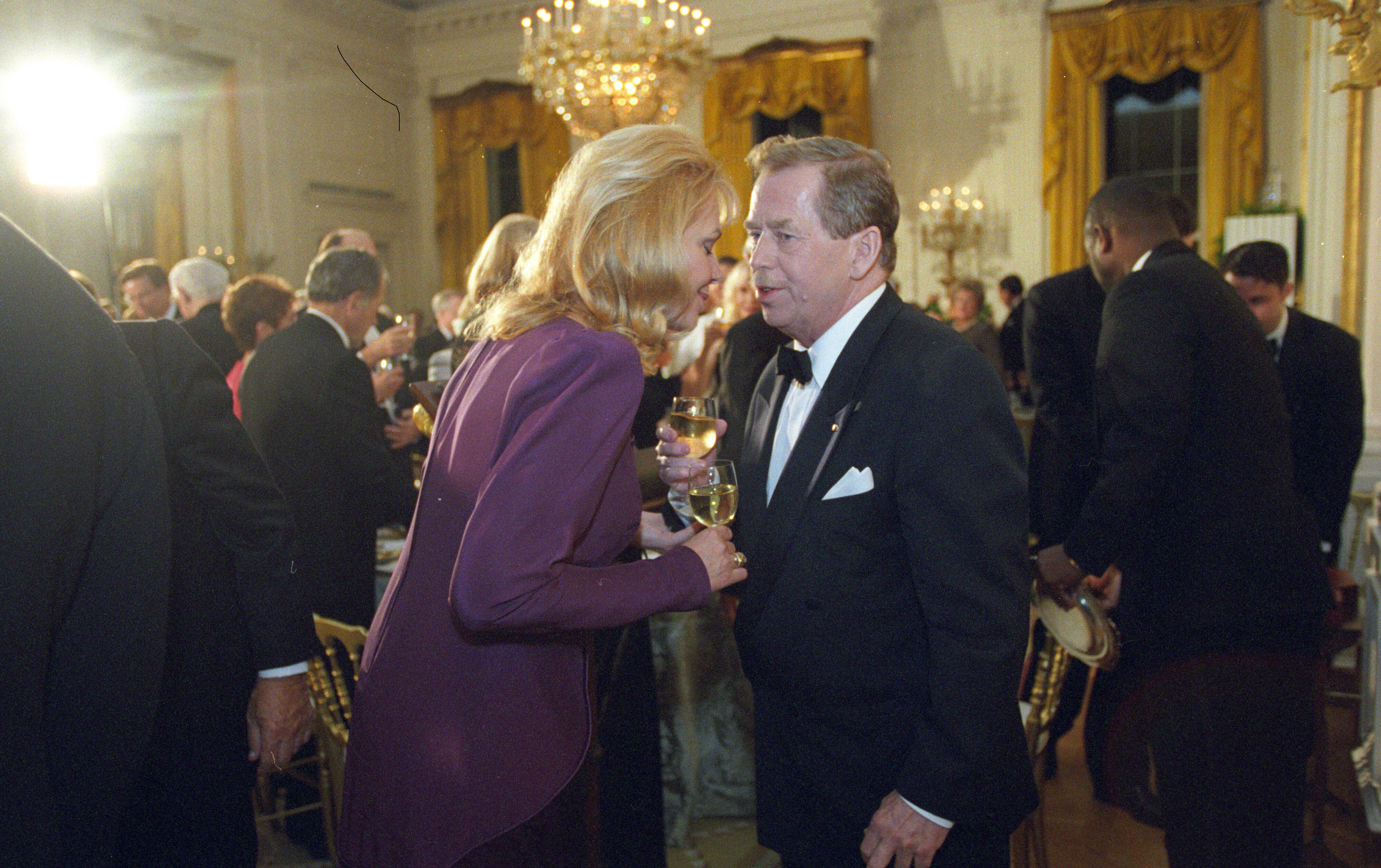 President Václav Havel holds a glass of sparkling wine in his hand and leans toward his wife, Dagmar Havlová. Havlová leans towards Havel and holds a glass of wine. Havel wears a tuxedo, and Havlová wears a violet/ or purple dress. 