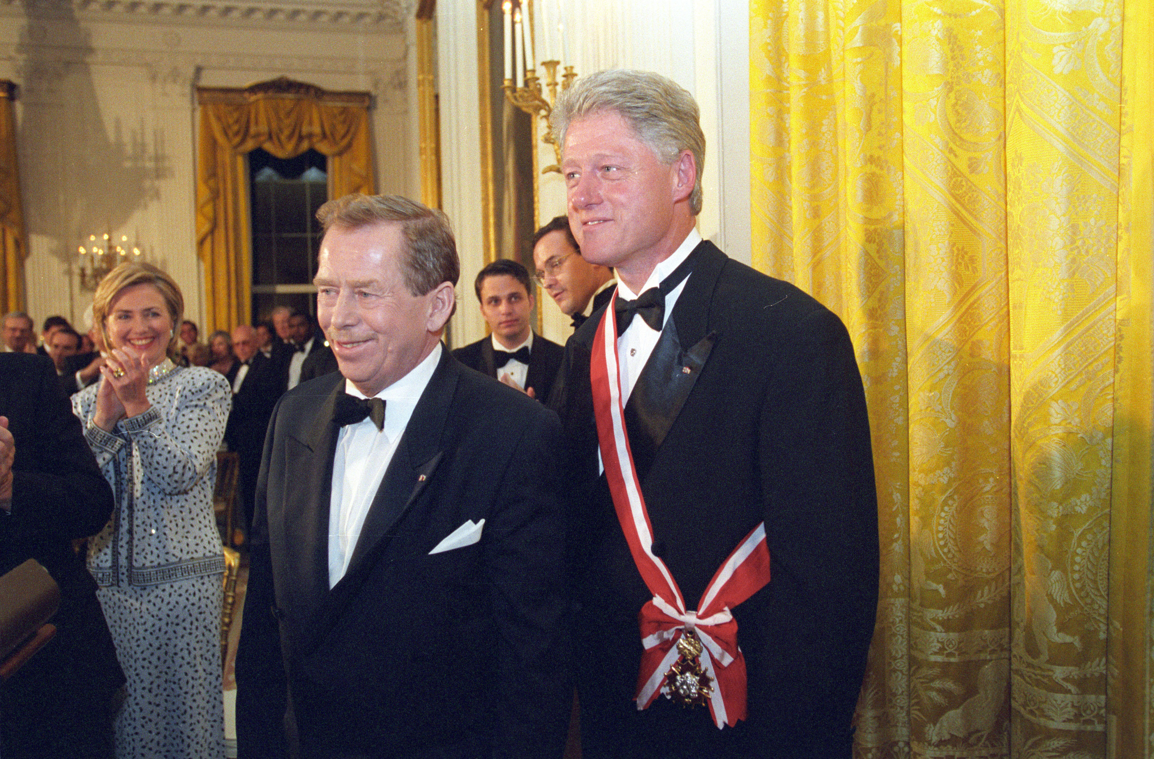 President Clinton wears a red and white sash over black tuxedo suit and white shirt.  President Clinton stands, posing for a photograph with President Václav Havel. First Lady Hillary Rodham Clinton stands, clapping and smilling behind the two presidents.
