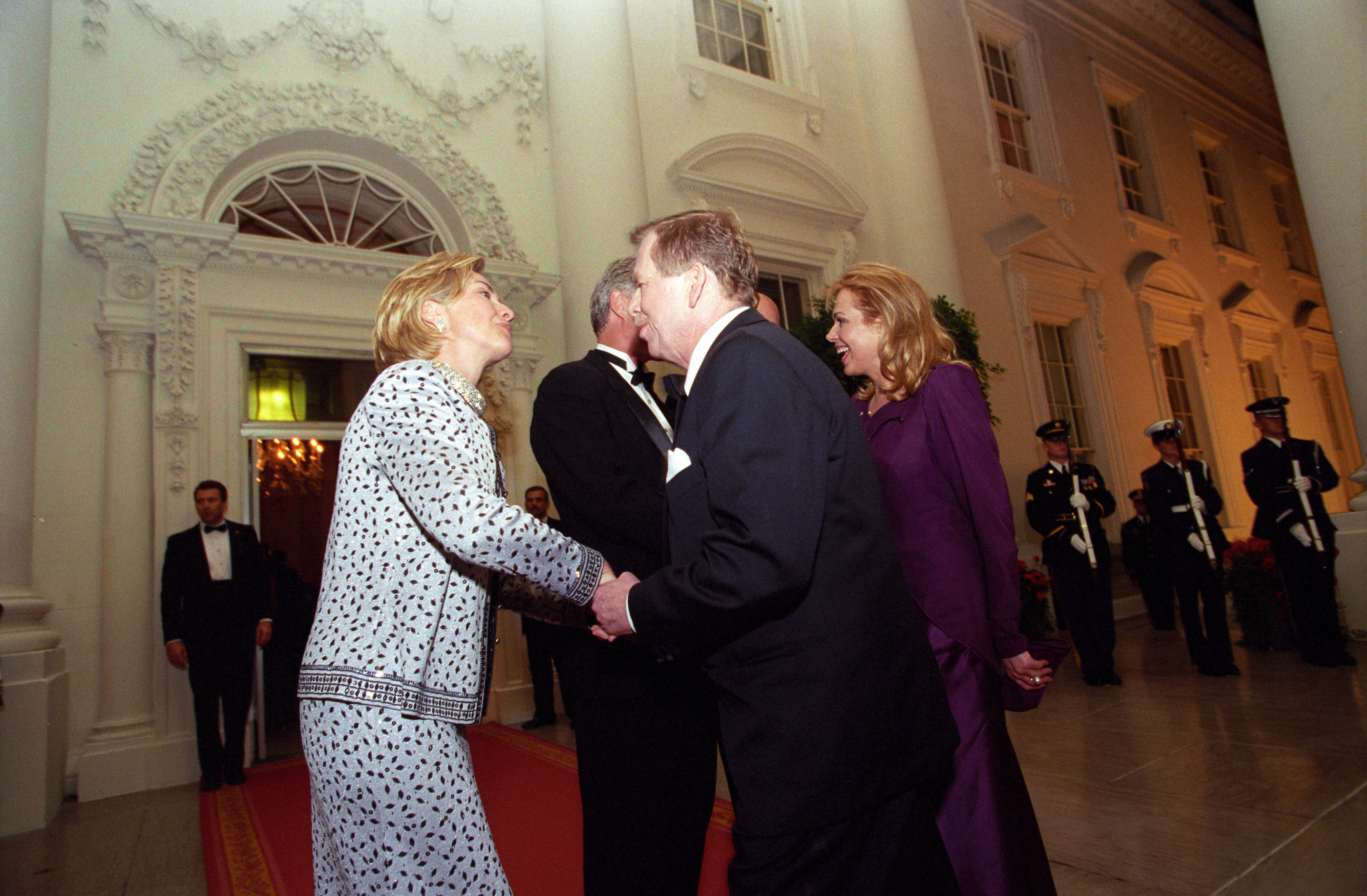 First Lady Hillary Rodham Clinton stands and shakes hands with President Václav Havel. President Clinton stands and speaks with Dagmar Havlová. They stand outside of the White Houise. Three uniformed soldiers can be seen in the background standing at attention, holding rifles in front of them.