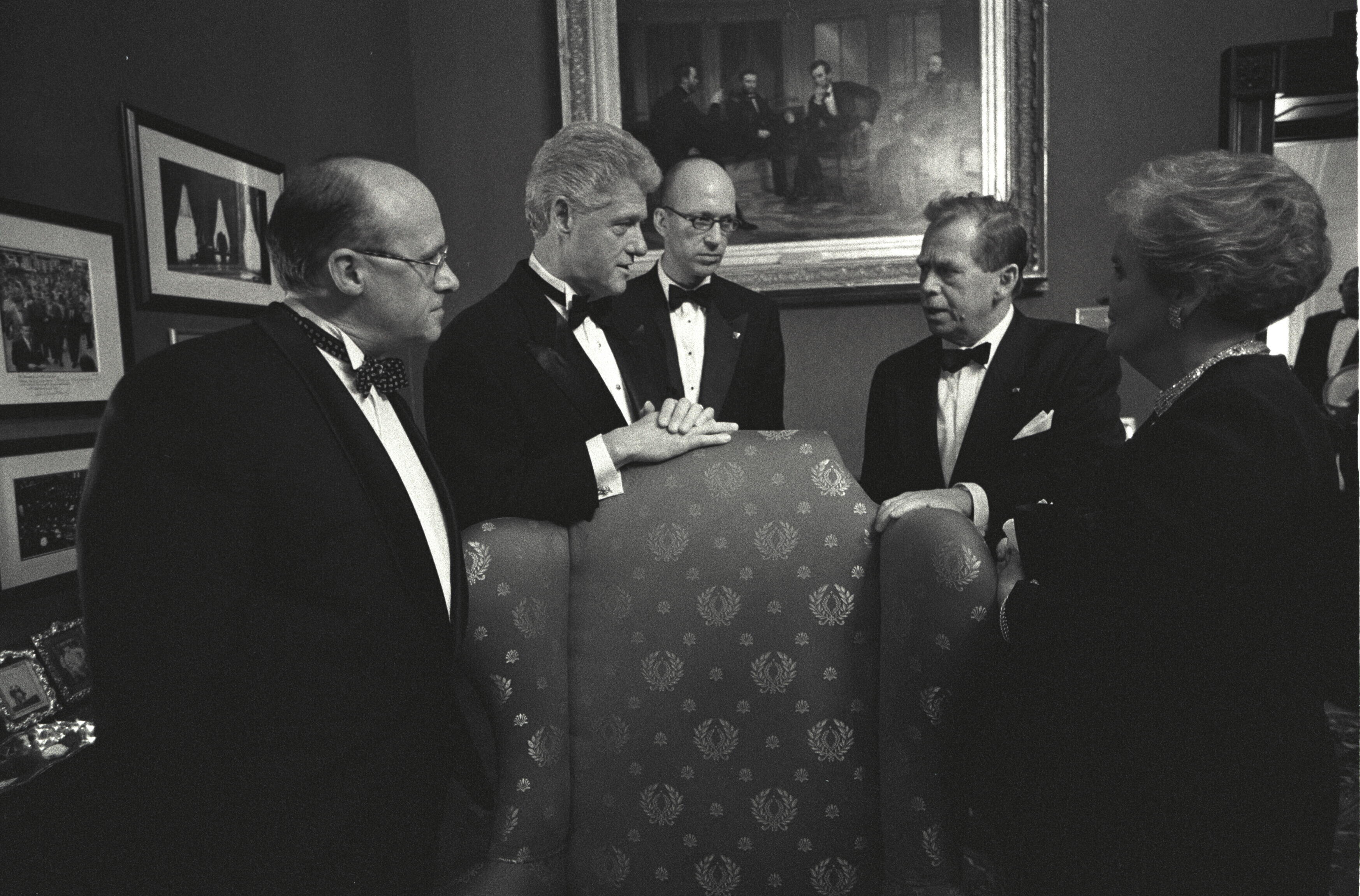 Black and white photograph. President Clinton stands and talks with President Václav Havel, Secretary Madeleine Albight, and two unidentified men stand in the White House Treaty Room. President Clinton places his hands folded on a wing pack chair. The men are dressed in tuxedos. The painting "The Peacemakers an 1868 painting hangs on the wall. 