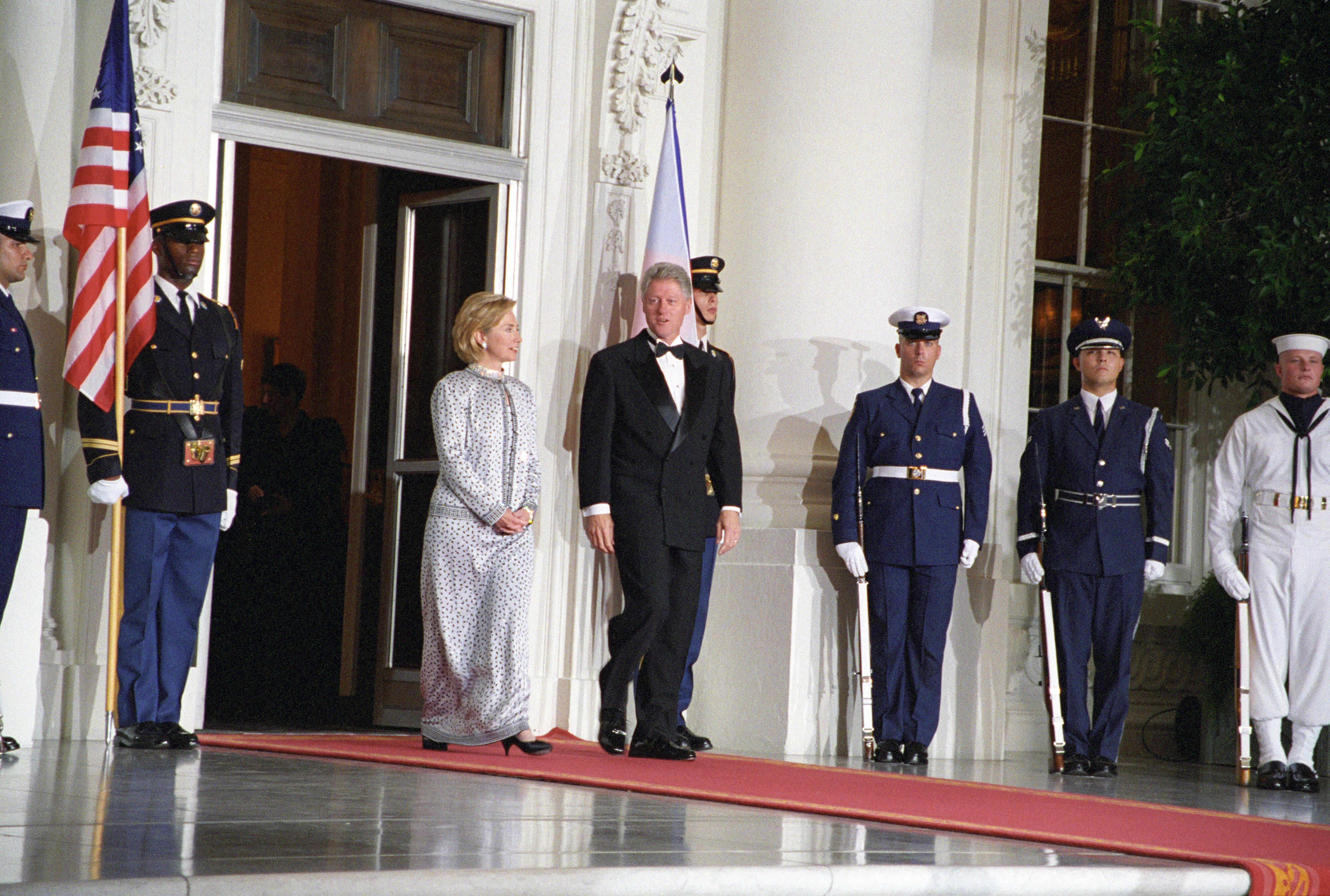President Clinton and First Lady Hillary Rodham Clinton stand omn a red carpet in front of the White House. Six uniformed military personnel stand at attention. 