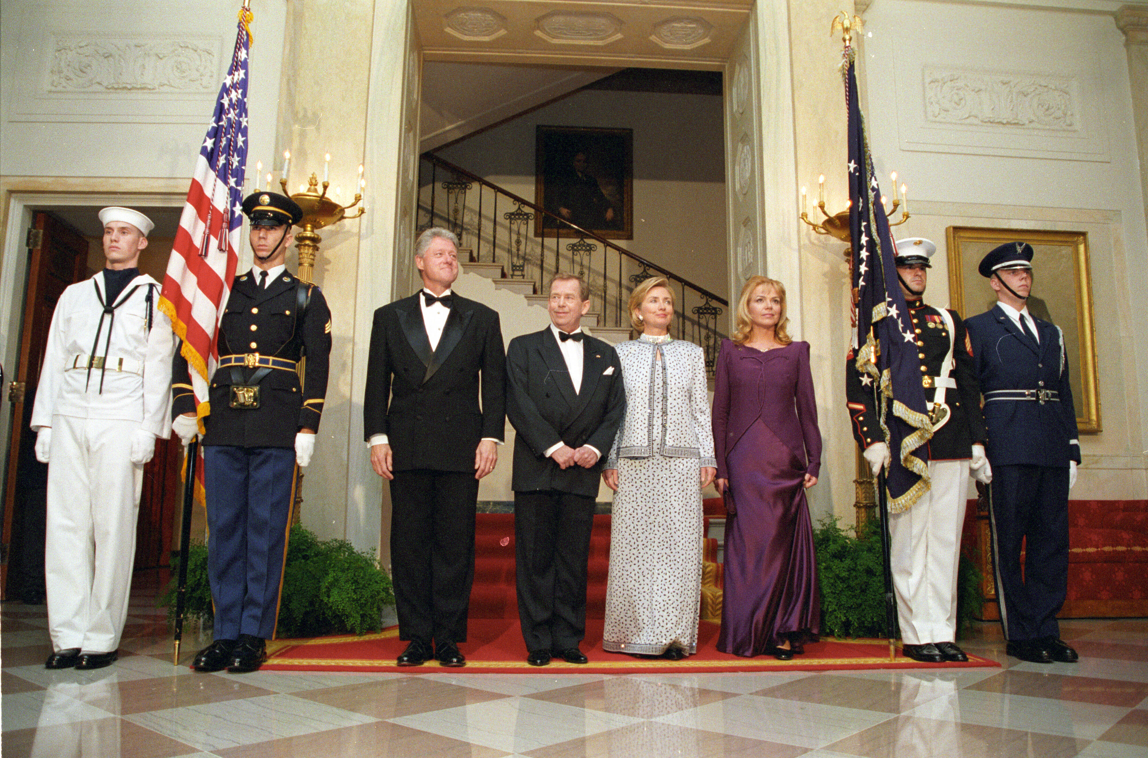 President Clinton, First Lady Hillary Rodham Clinton, President Václav Havel, and Dagmar Havlová stand on a red carpet on a tiled floor. Four uniformed military personnel stand at attention. A United States Flag and the flag of the President stand in the background.