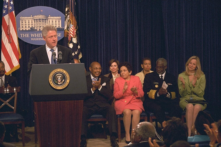 President Clinton stands behind a podium. people sit behind the President, clapping at his remarks.