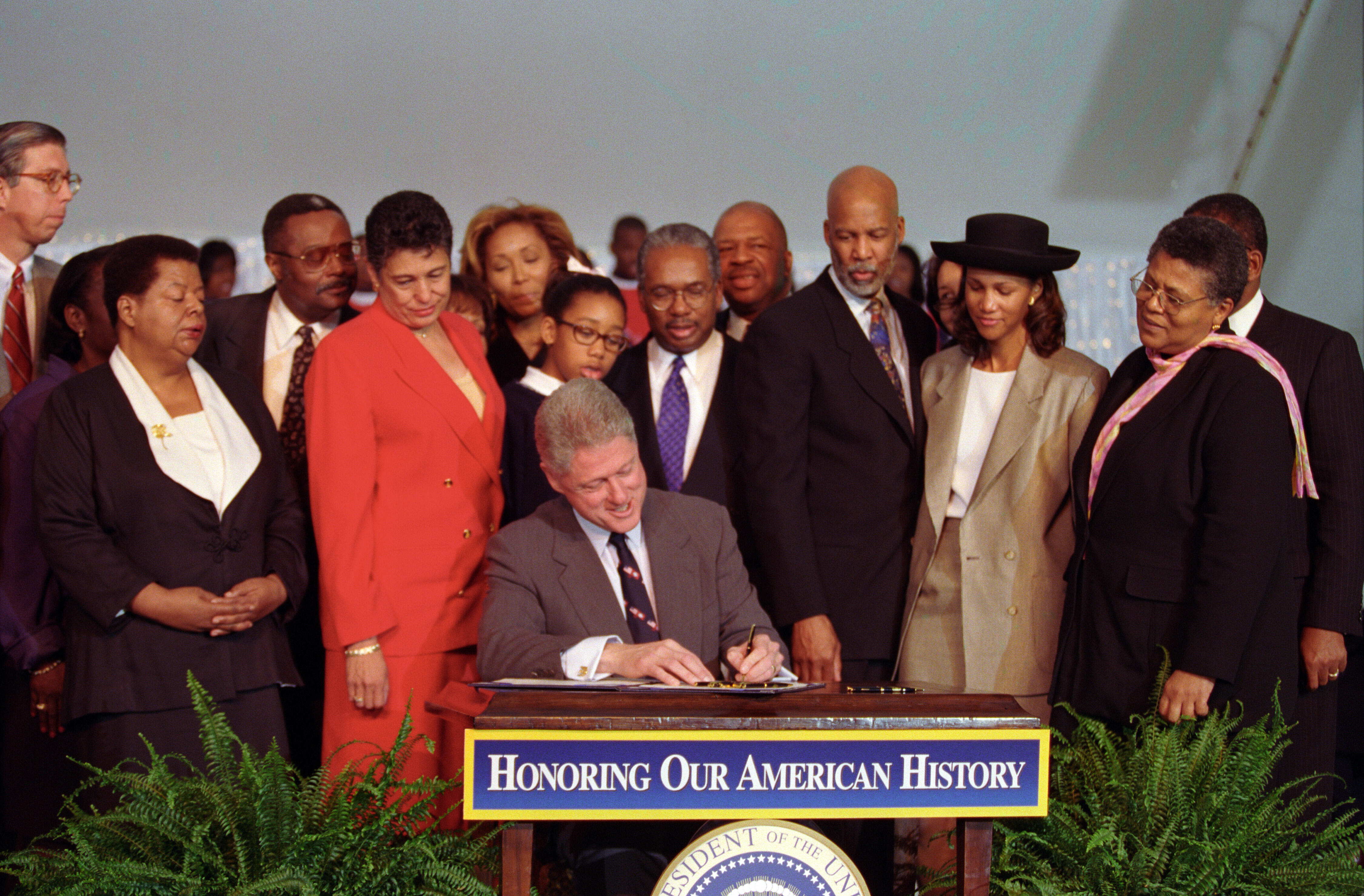 President Clinton sits at a desk. The Little Rock Nine and their families stand, looking down at President Clinton while he signs the Little Rock Central High School National Historic Site Bill.