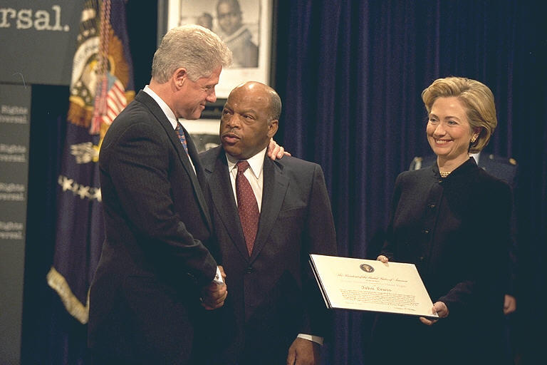 President Clinton and Hillary Rodham Clinton presents Rep. John Lewis with the Eleanor Roosevelt Award for Human Rights. Hillary Rodham Clinton holds the award in her hands. President Clinton stands with his left hand on Rep. Lewis's shoulder while shaking Lewis's hand with his right hand. 