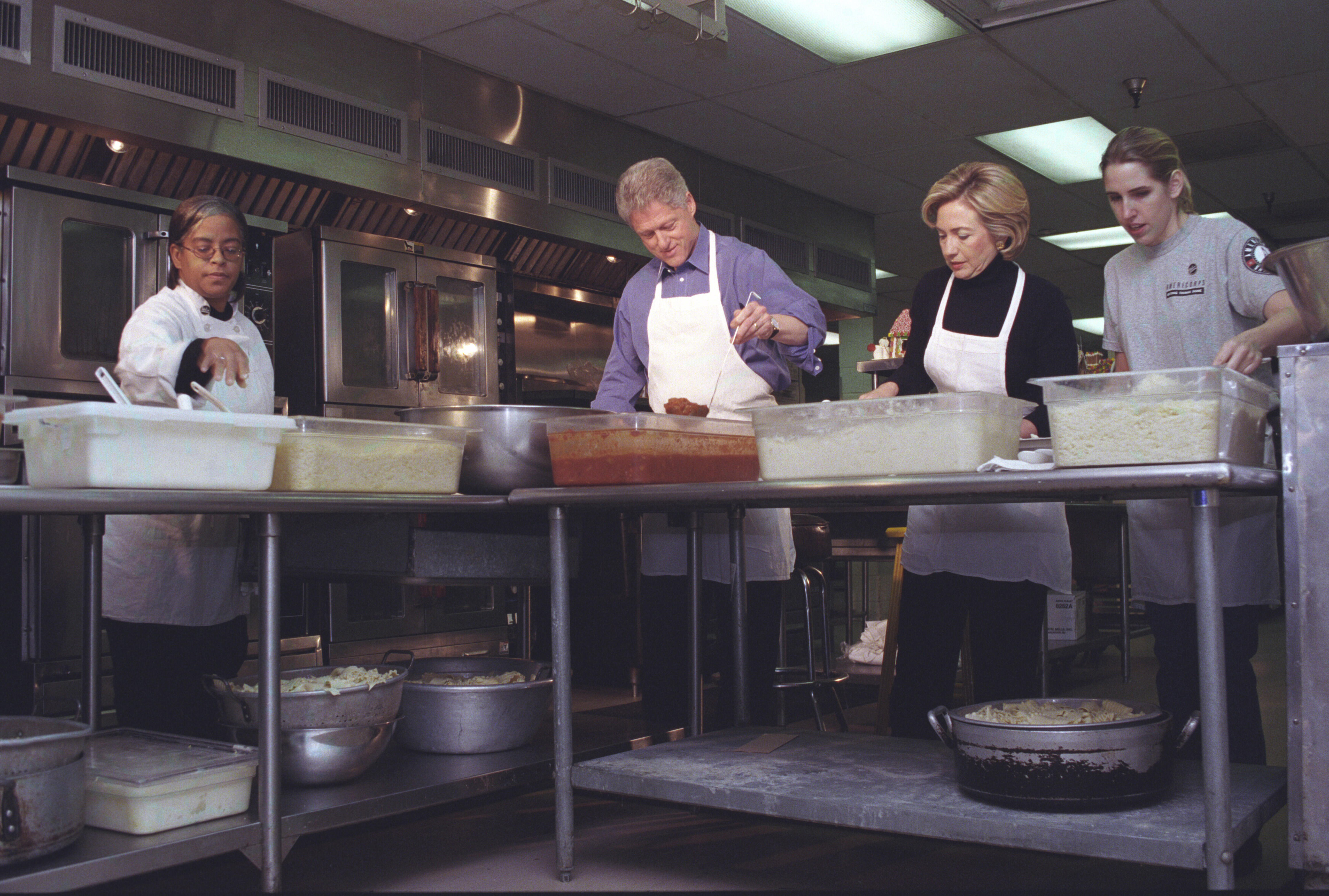 President Clinton and First Lady Hillary Rodham Clinton stand, wearing white aprons, while preparing food in a kitchen. Two other volunteers assist the Clintons.