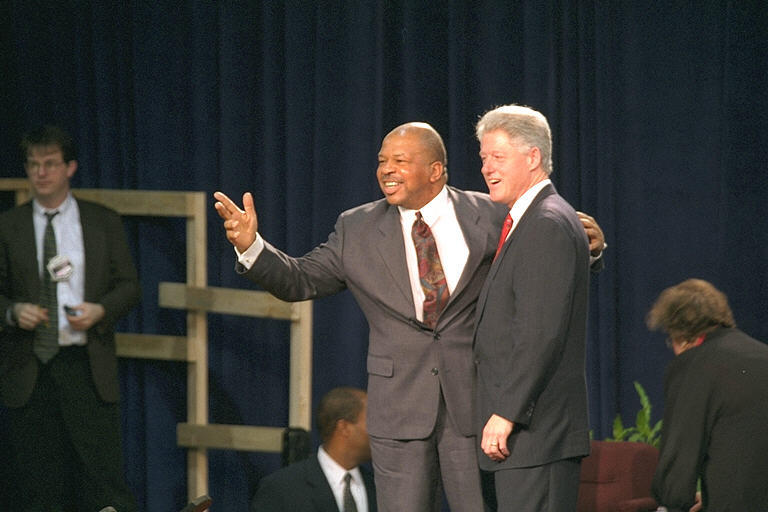 President Clinton stands on a stage with Rep. Elijah Cummings. Cummings wraps his left arm around President Clinton's back. Cummins extends his right arm out toward the crowd.