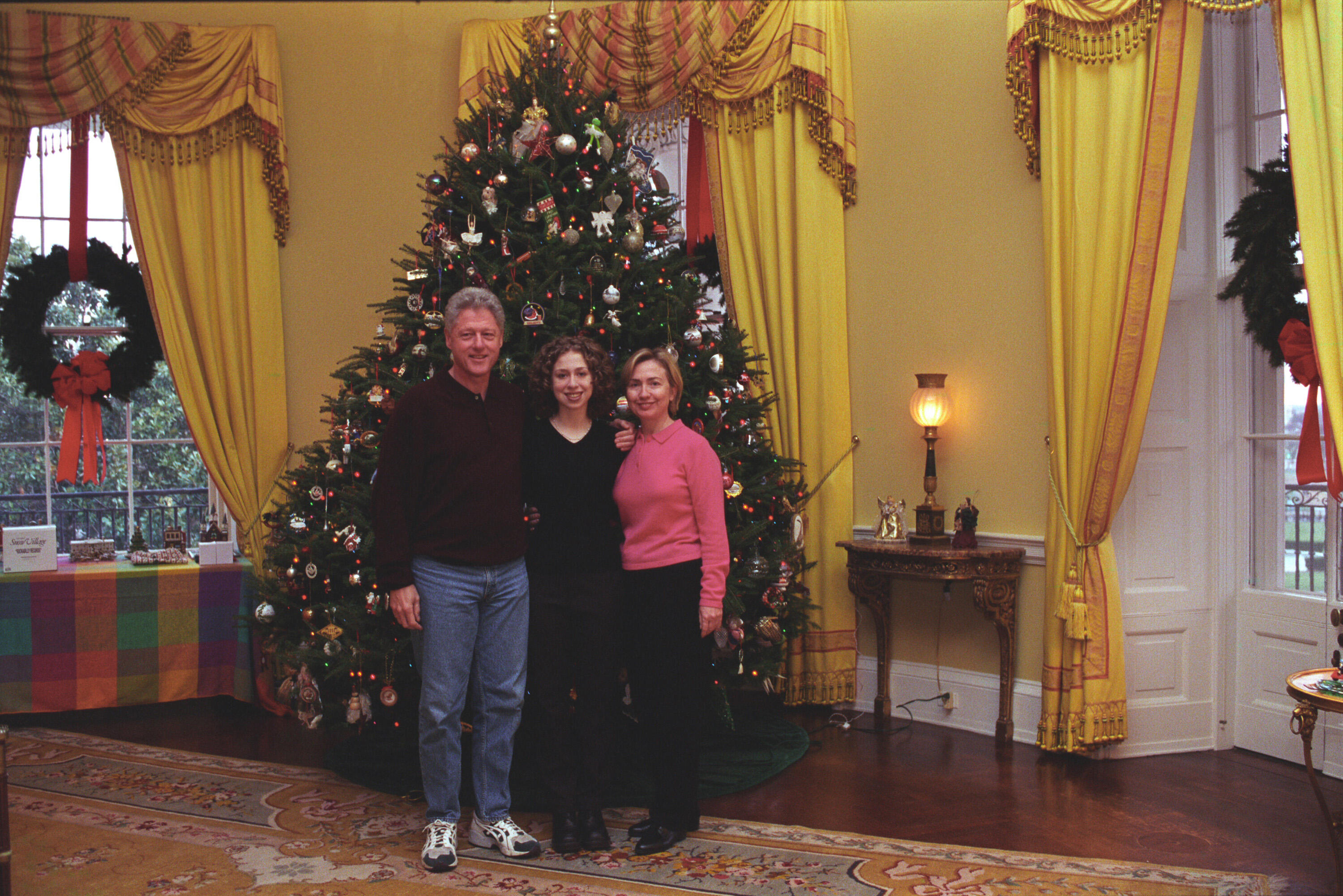 President Clinton, Hillary Rodham Clinton, and Chelsea Clinton pose together in front of the family Christmas tree in the Yellow Oval Room in the White House. The First Family is dressed in casual dress. The President wears jeans and sneakers.