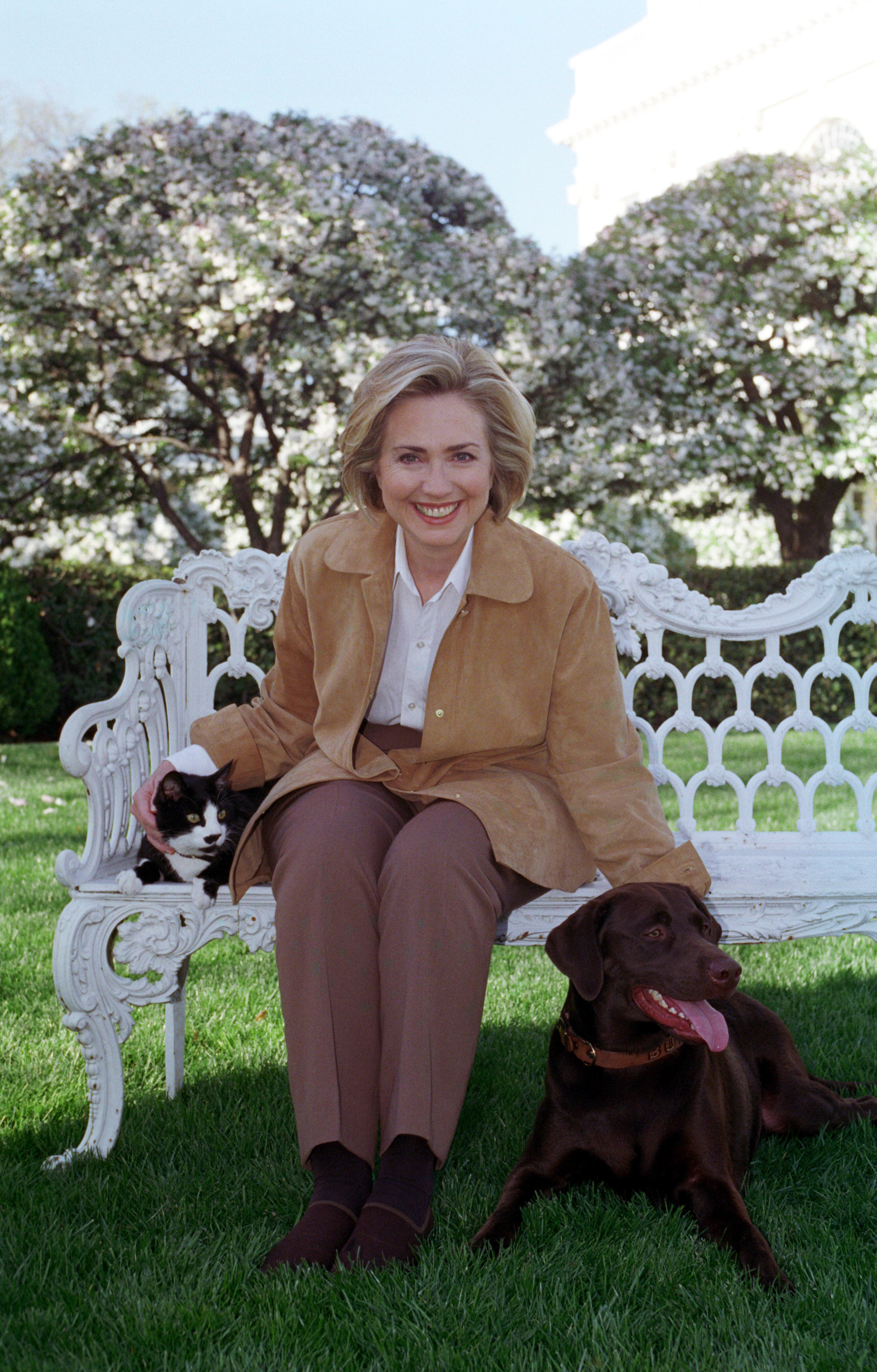 First Lady Hillary Rodham Clinton sits on a white metal bench with Socks the cat on the South Lawn of the White House. She pets Buddy the dog that is laying at her feet