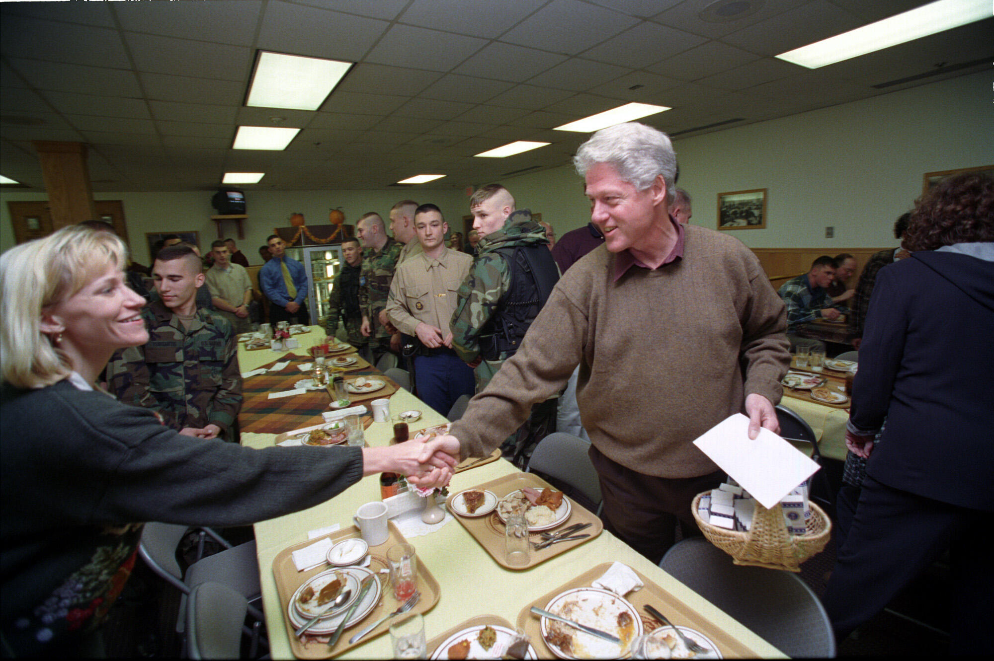 President Clinton stands and shakes a woman's hand while standing over a long table. The table contains plates of food. Military personnel and their families stand in the background.
