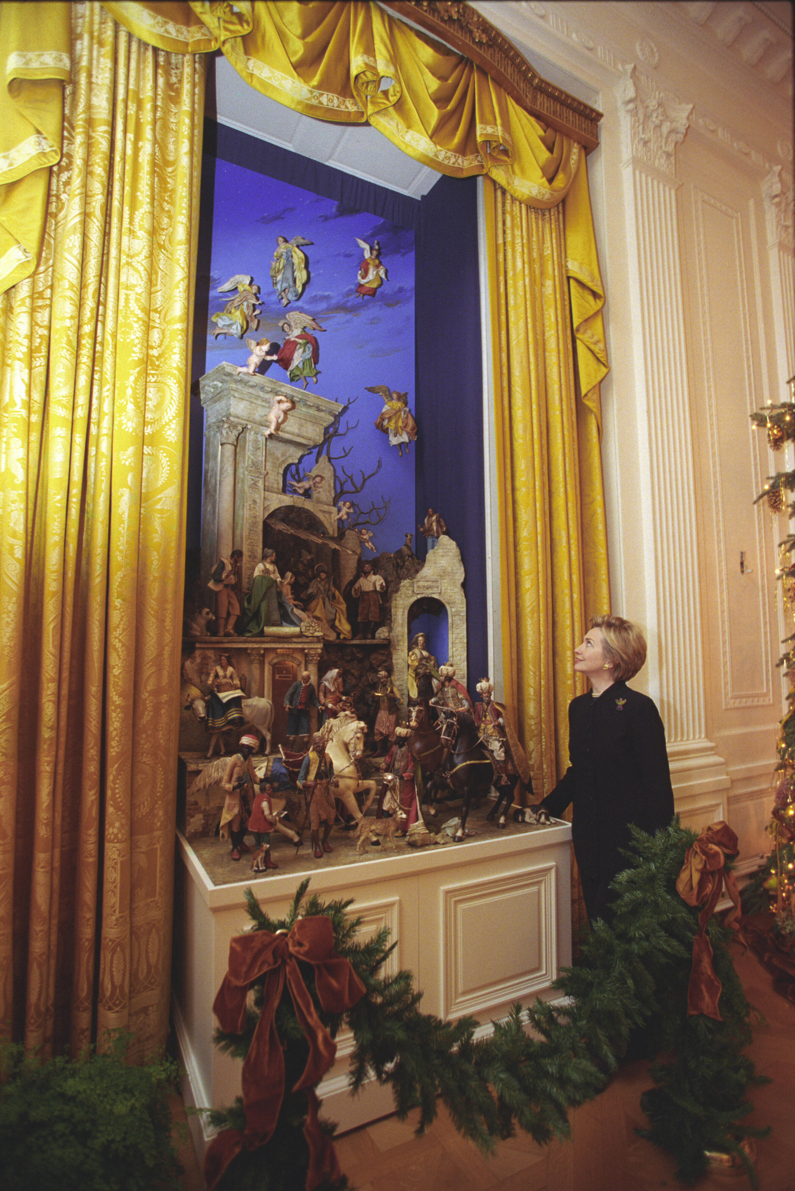 First Lady Hillary Rodham Clinton looks at a creche. The creche is surrounded by a yellow curtain.