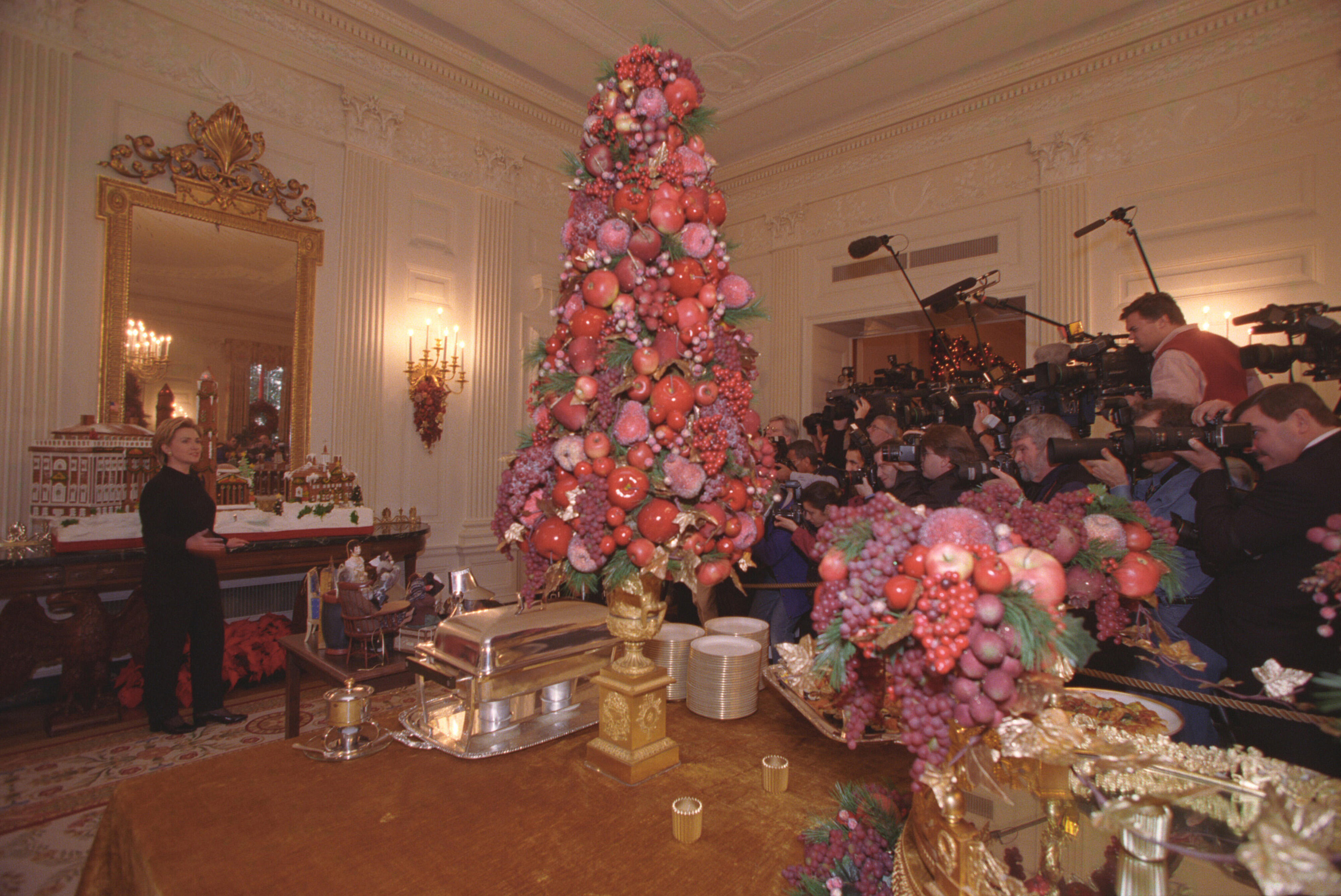 First Lady Hillary Rodham Clinton stands in a room with a Christmas tree made of imitation fruit. The tree and other decorations in the room are pink, violet, and green. A  group of press point cameras and microphones toward the First Lady.