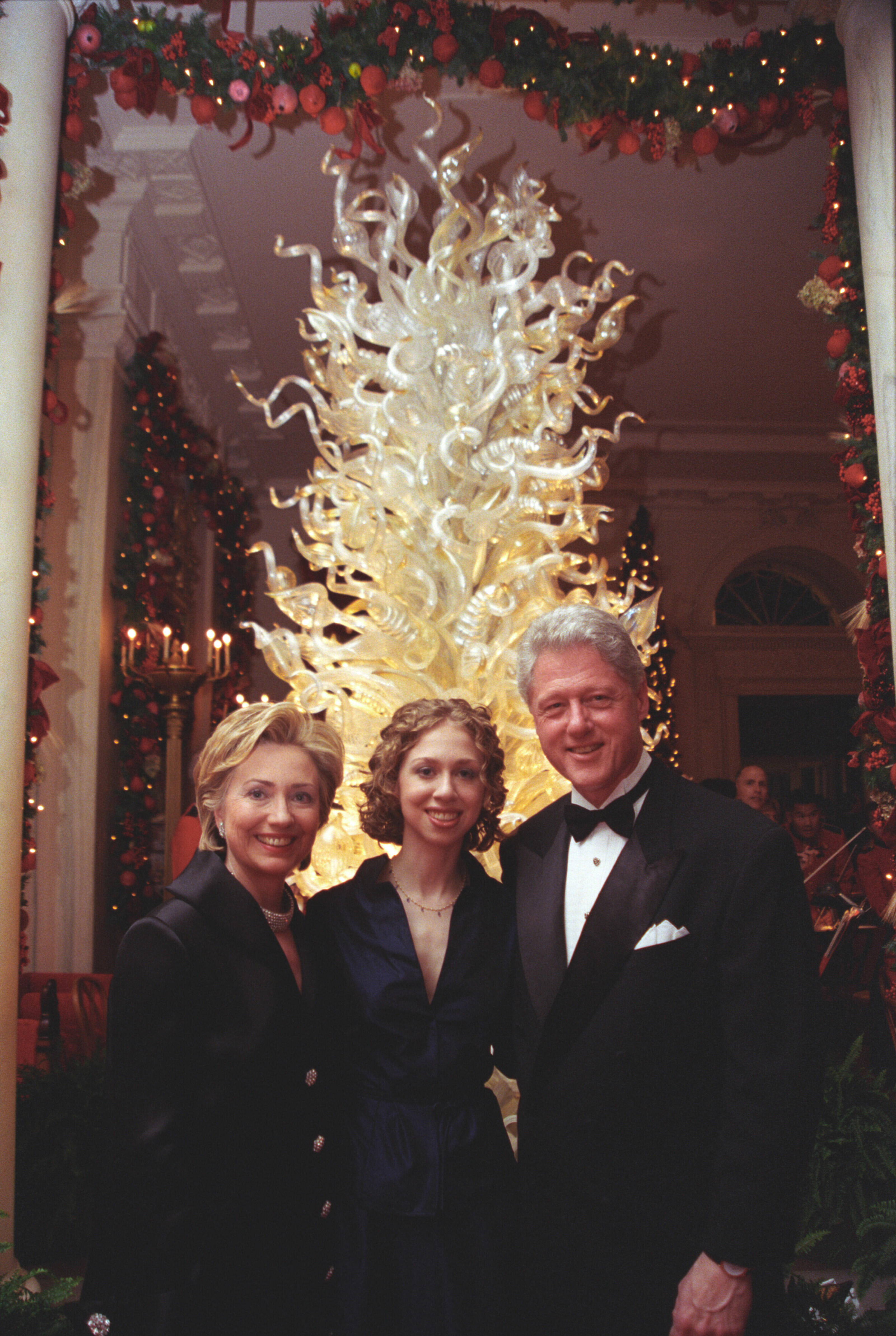 President Clinton, Hillary Rodham Clinton, and Chelsea Clinton stand in front of a Dale Chihuly Christmas Tree in the Grand Foyer of the White House. The tree is lit brightly. 