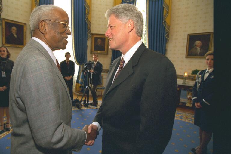 President Clinton stands shanking ands and looking directly into the eyes of John Hope Franklin. Franking wears glasses. Both men wear suits. 4 other people can be seen standing in the background of the photograph.