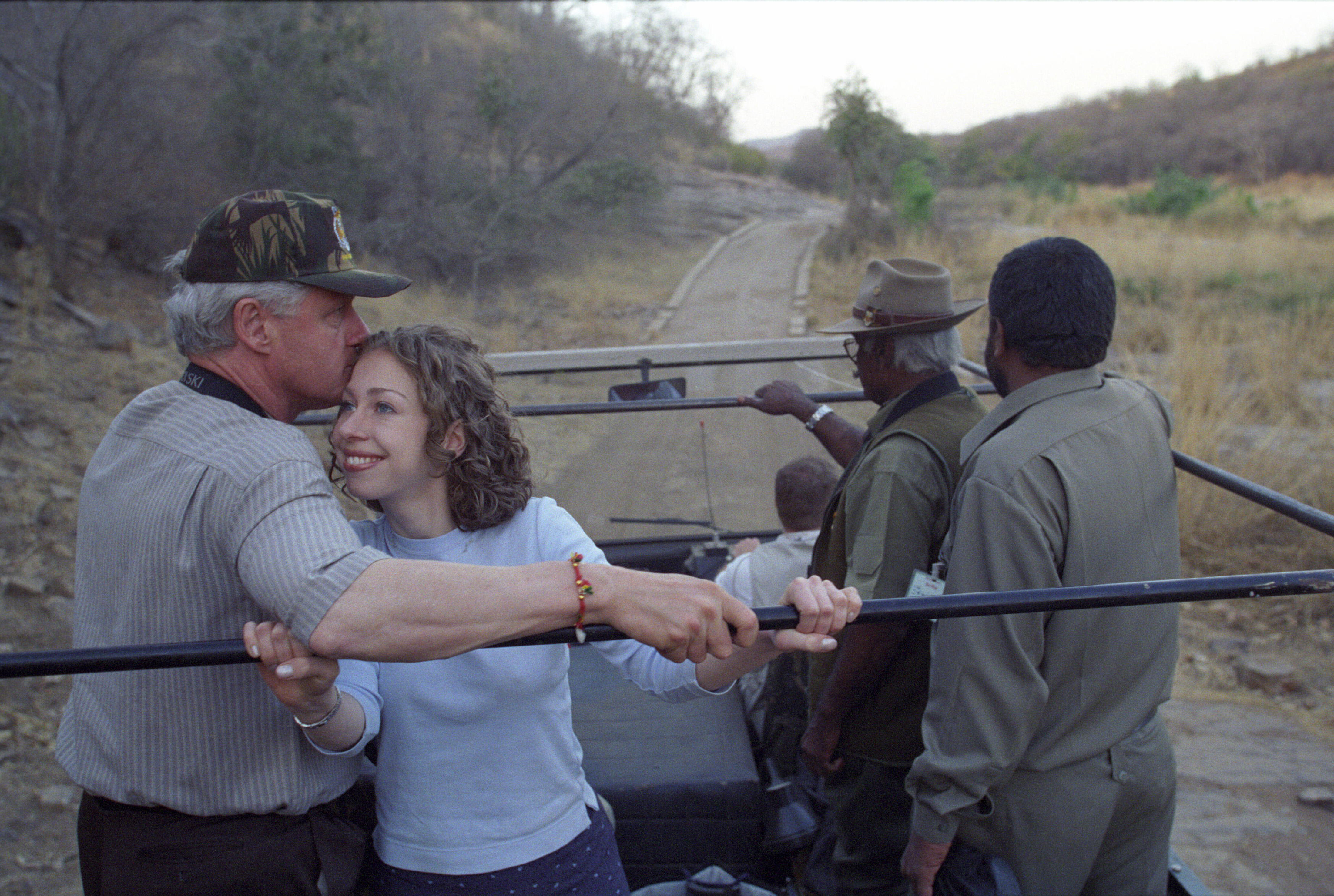 President Clinton kisses Chelsea Clinton's head while standing in the back of a jeep. They both hold rails in the jeep as they stand. Two other men can bee seen standing in the back of the jeep. 