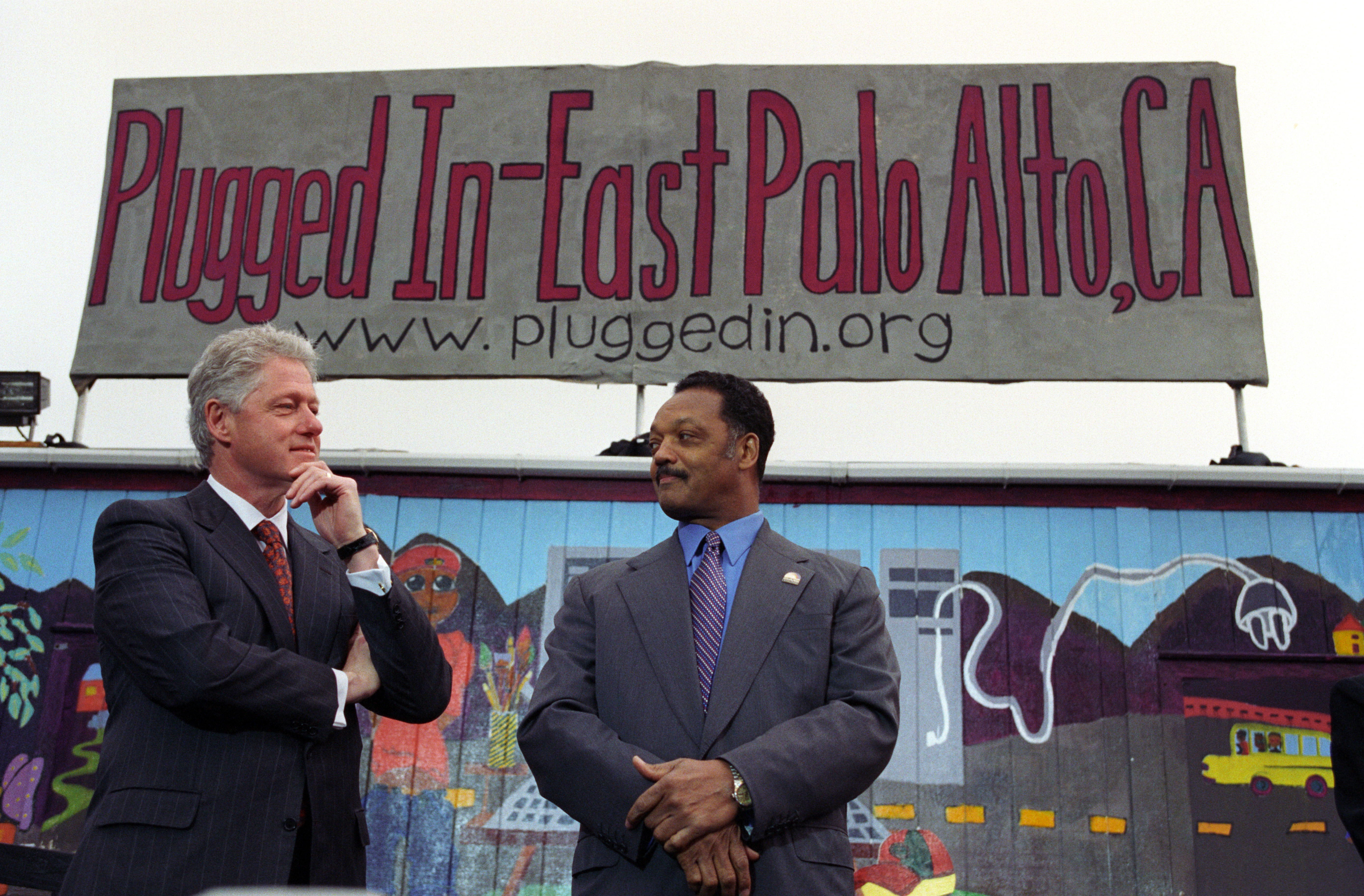 President Clinton and Rev. Jesse Jackson stand in front of a sign that reads "Plugged In-East Palo Alto, CA." President Clinton holds his hand under his chin. Jackson crosses his hands in front of him.