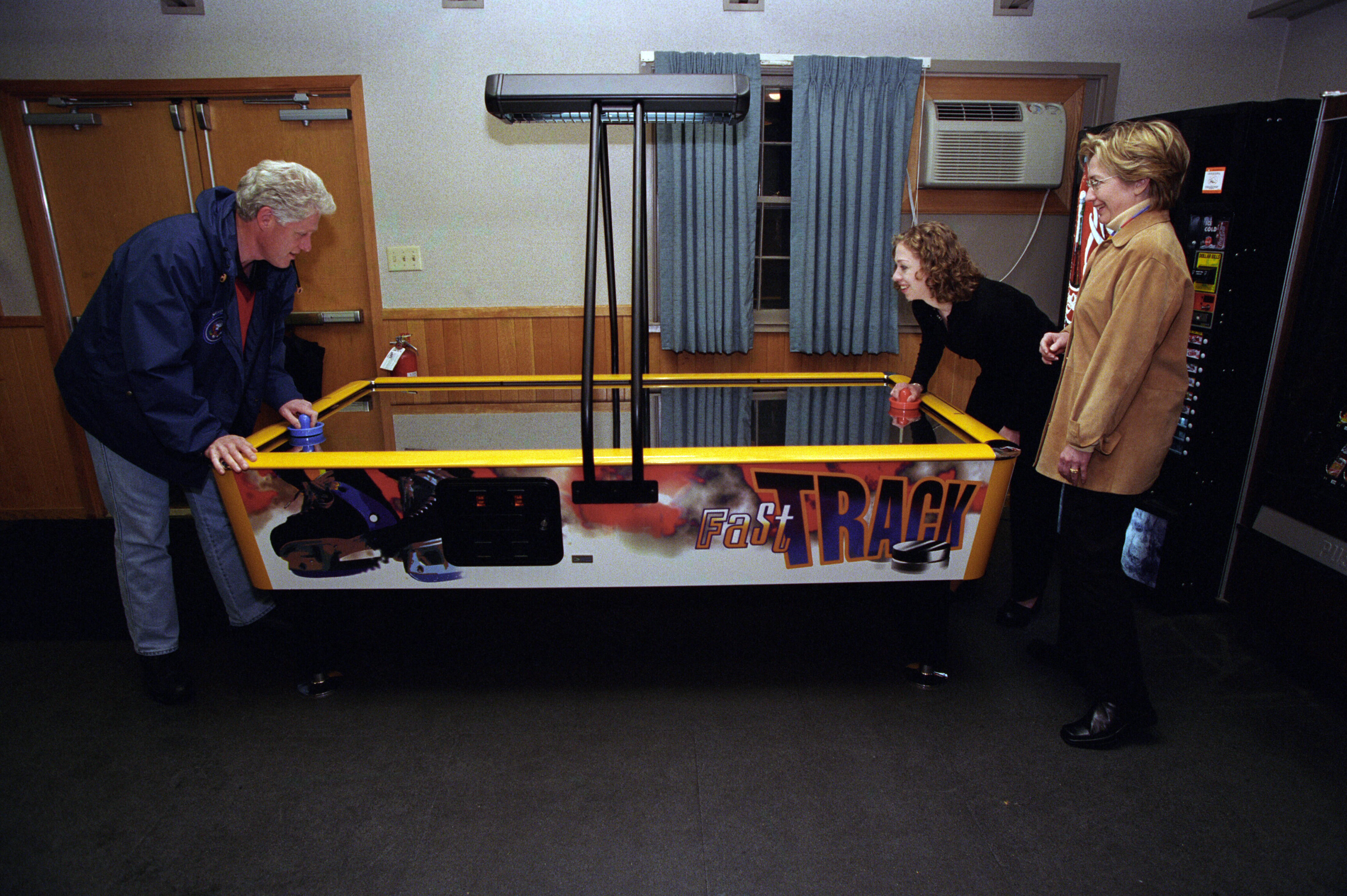 Hillary Rodham Clinton stands and watches President Clinton and Chelsea Clinton play air hockey. They all stand. The President and Chelsea Clinton lean over the air hockey table as they play. 