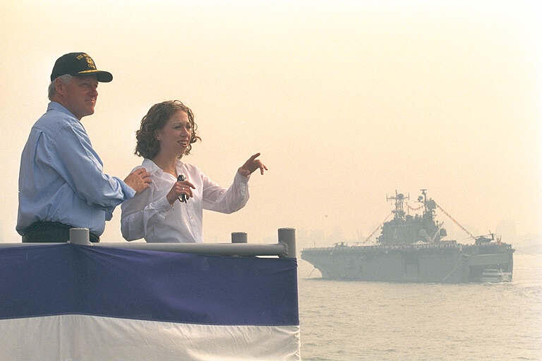 President Clinton and daughter Chelsea Clinton stand on a dock in the New York Harbor. Chelsea points at U.S. and International warships in the harbor. President Clinton wears a baseball cap.