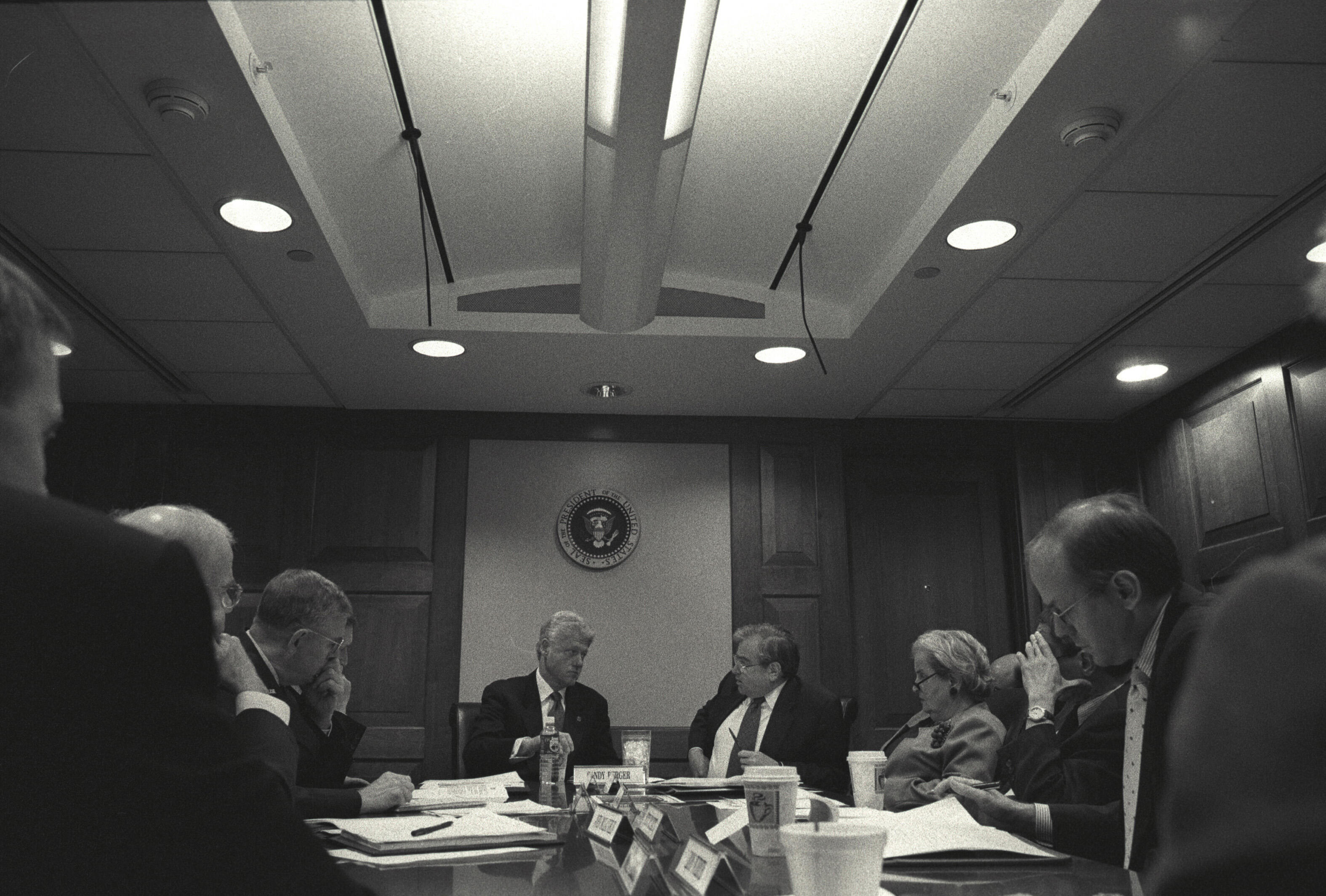 Black and white photograph of President William Jefferson Clinton in the Situation Room at the White House. Also in attendance are Secretary of State Madeleine Korbel Albright, National Security Advisor Samuel "Sandy" Berger, Secretary of Defense William Sebastian Cohen, and White House Chief of Staff John David Podesta