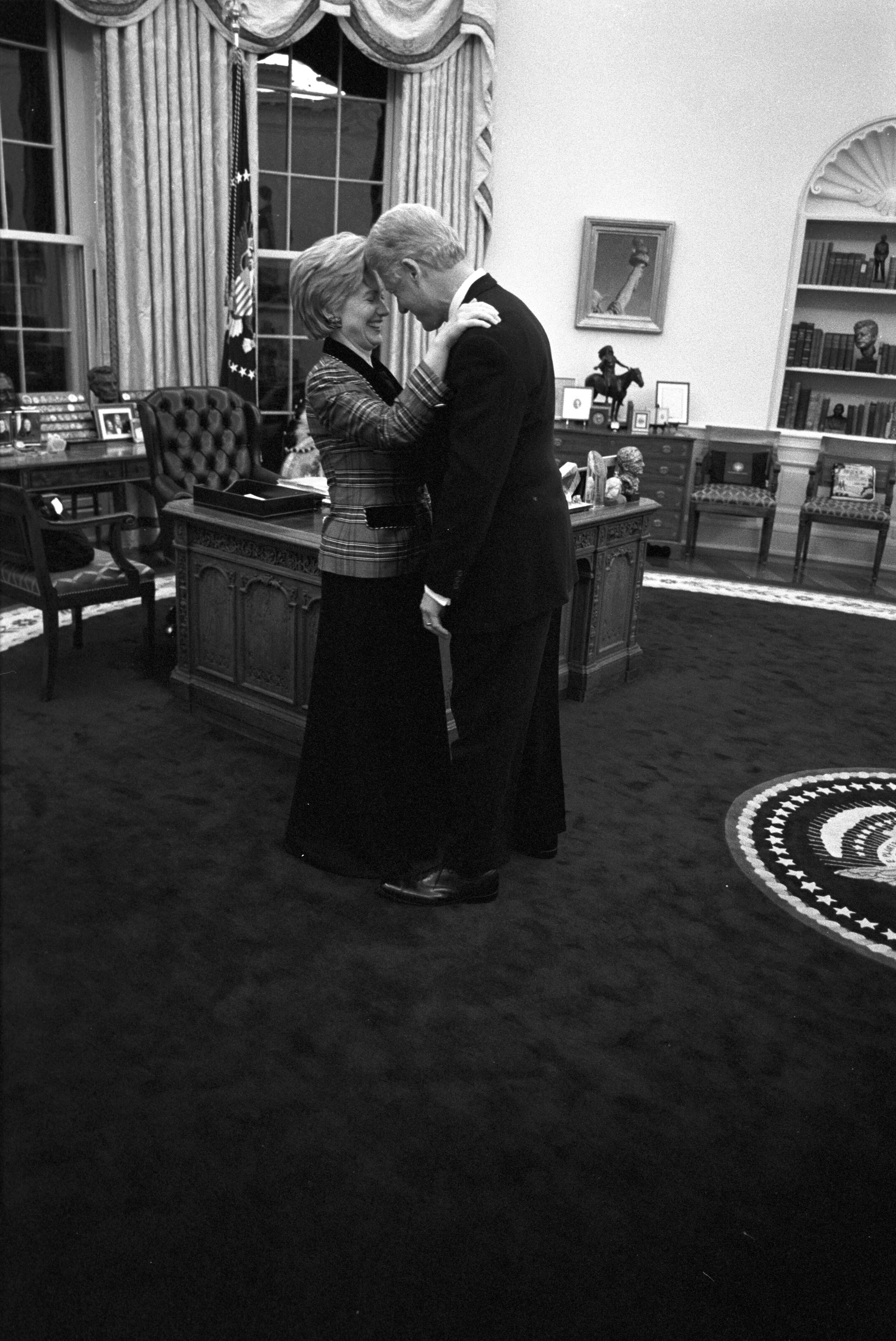 President Clinton and First Lady Hillary Rodham Clinton are alone in the Oval Office near the Resolute Desk. They stand close together with foreheads touching